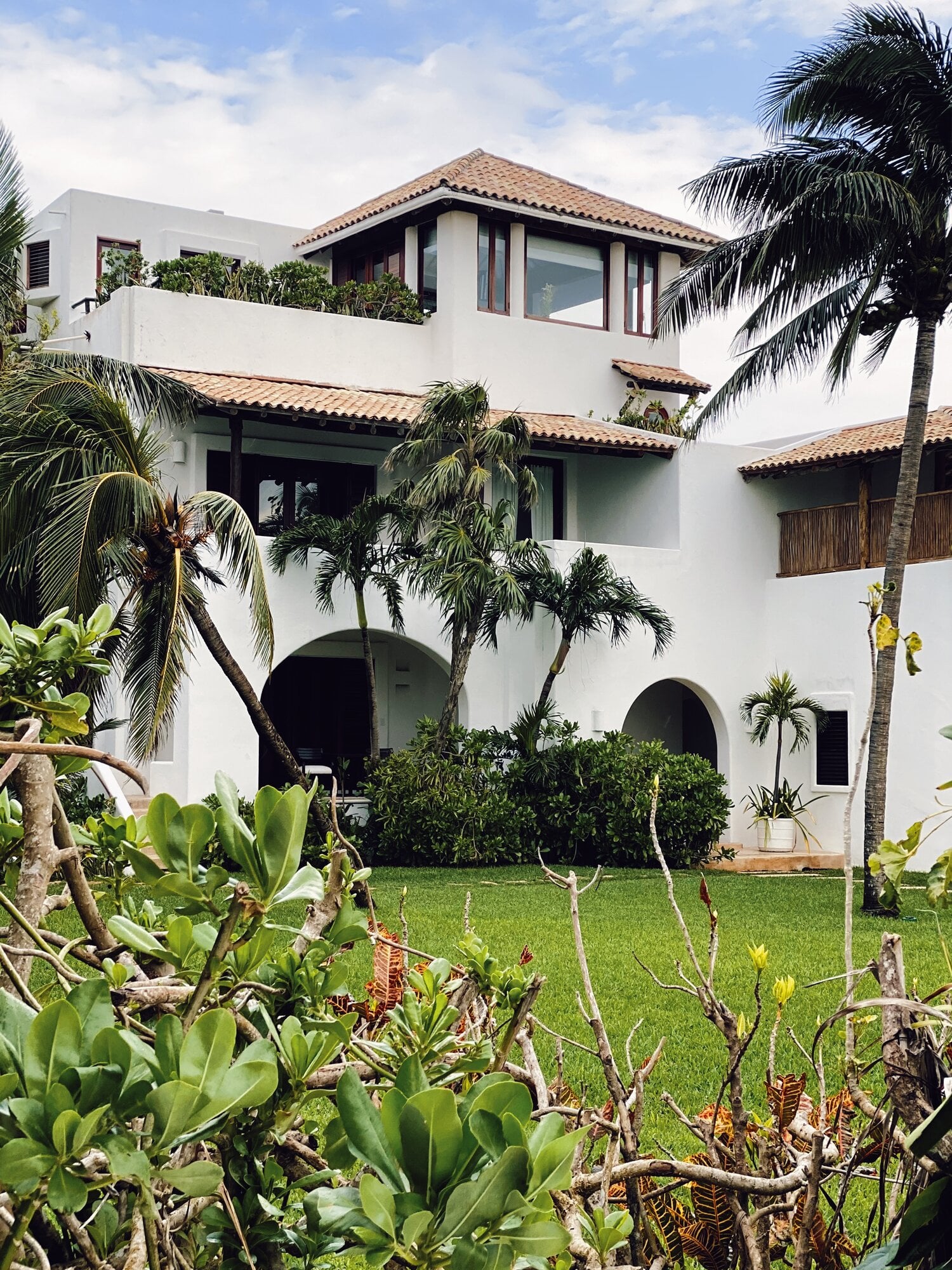 Hotel Esencia, Mexico — White stucco residence with terracotta roof tiles, arched openings, manicured lawn, palm trees, and tropical coastal modernist architecture.
