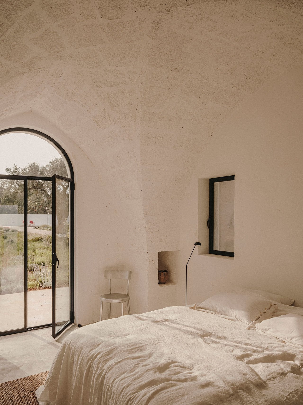 Borgo Gallana, Europe — Minimalist bedroom with arched window, concrete walls, linen bedding, and view of rural landscape through black-framed door.