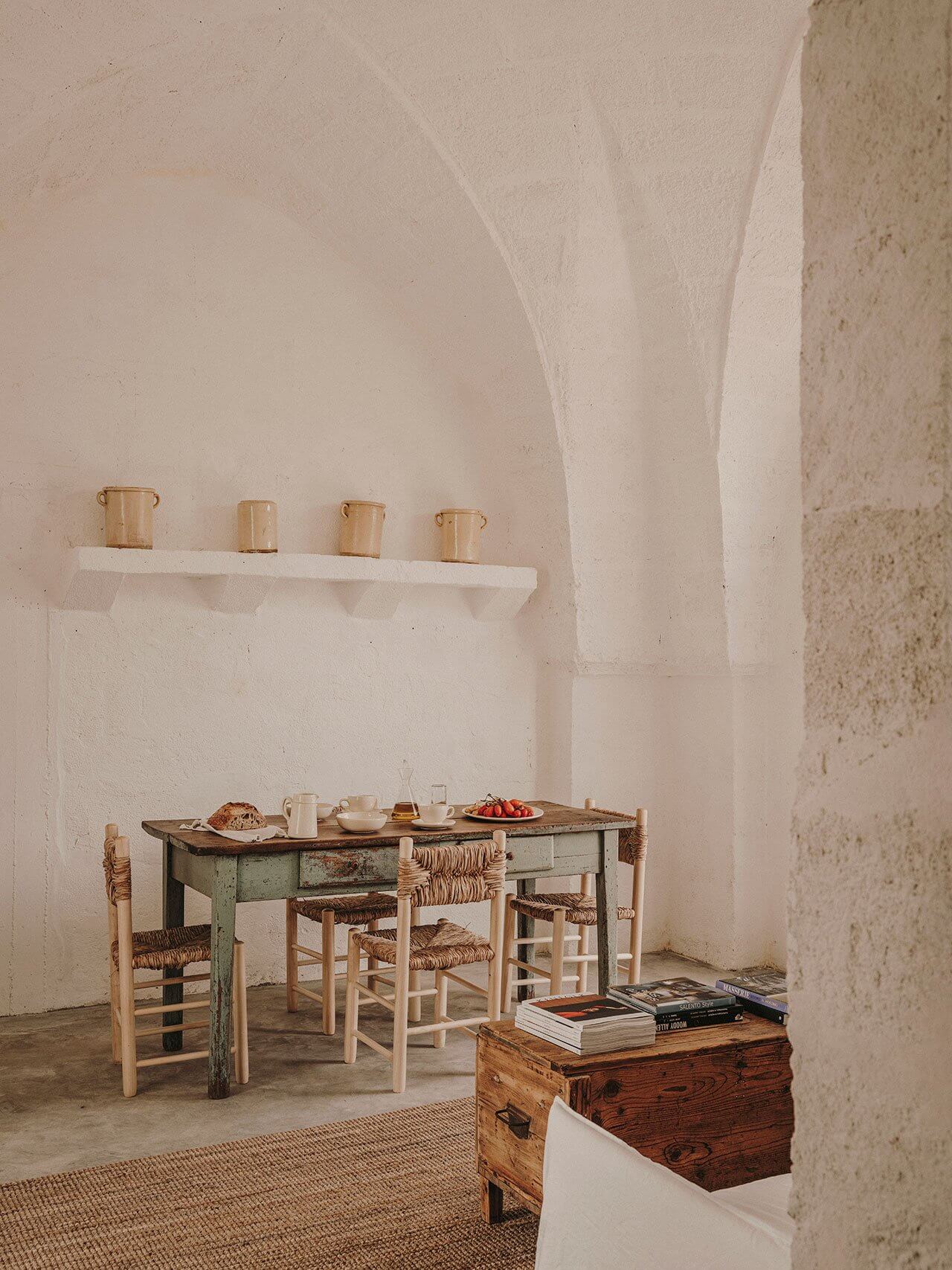 Borgo Gallana, Europe — Minimalist dining room with whitewashed walls, rustic wooden table, woven rush chairs, and vintage wooden trunk, sunlit Mediterranean interior.