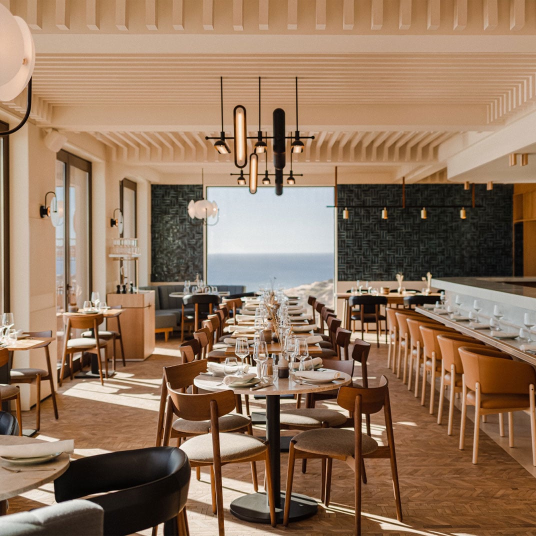 Aethos Ericeira, Portugal — Coastal dining room with wooden mid-century chairs, ocean views, geometric pendant lights, and ribbed ceiling detail.