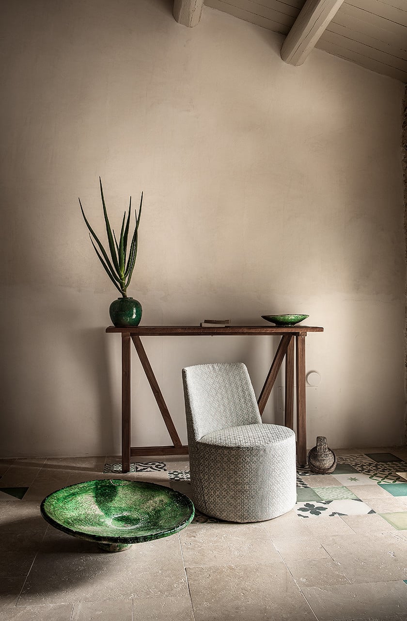 Dimora delle Balze, Europe — Minimalist interior with linen chair, wooden console table, green ceramic vessels, and patterned tile floor beneath exposed beam ceiling.