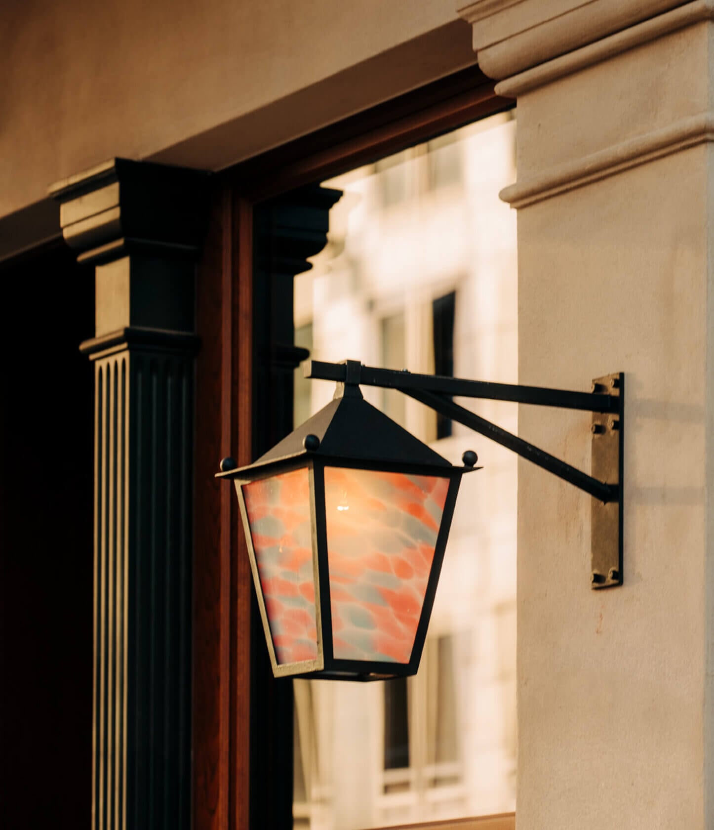 Château Royal, Europe — Wrought-iron wall sconce with amber glass panels casting warm light on cream stone facade at dusk, classical hospitality architecture detail.