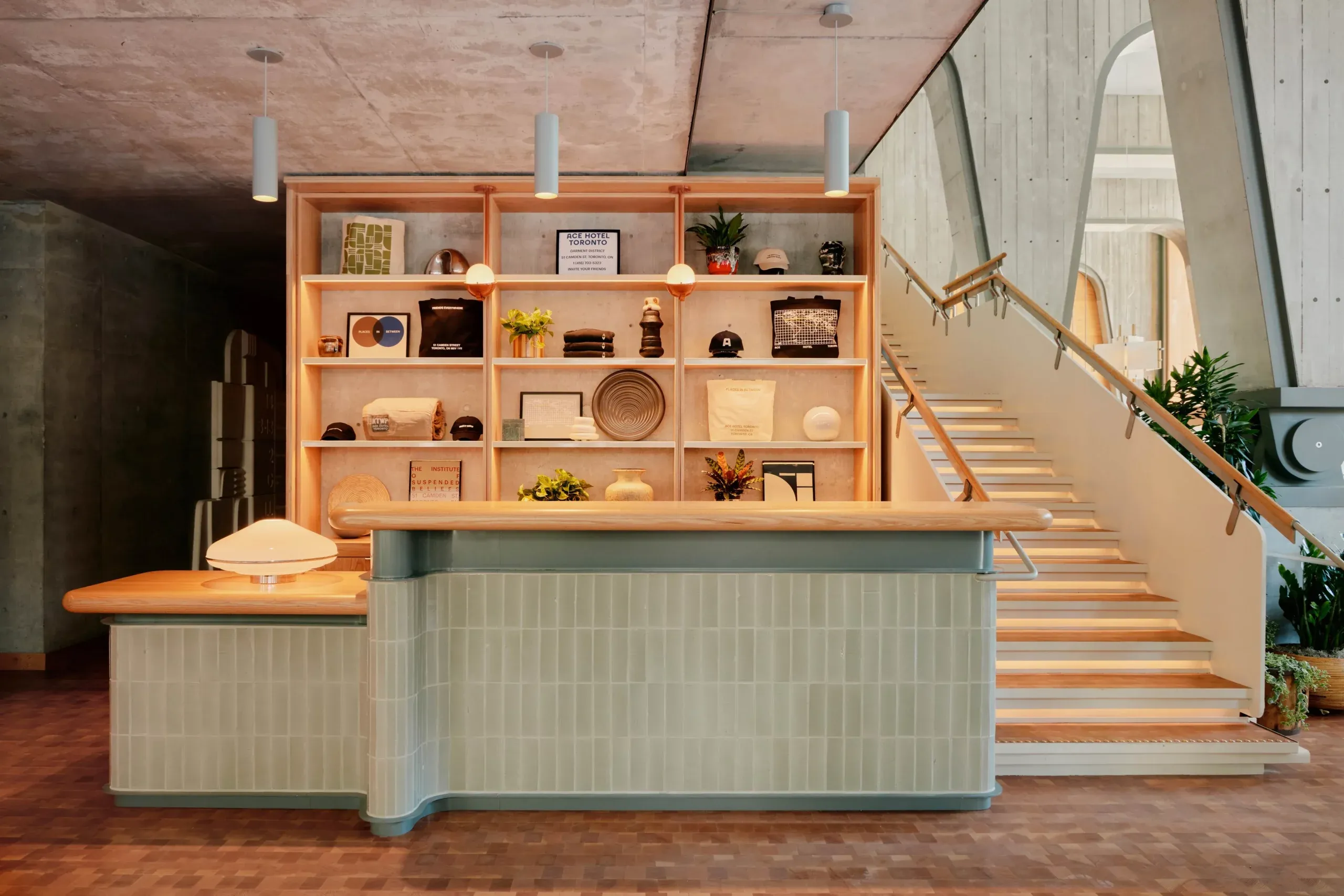 Ace Hotel Toronto, Canada — Minimalist reception desk with sage-green tiled front, warm wood shelving, pendant lights, and concrete ceiling in modern lobby interior.