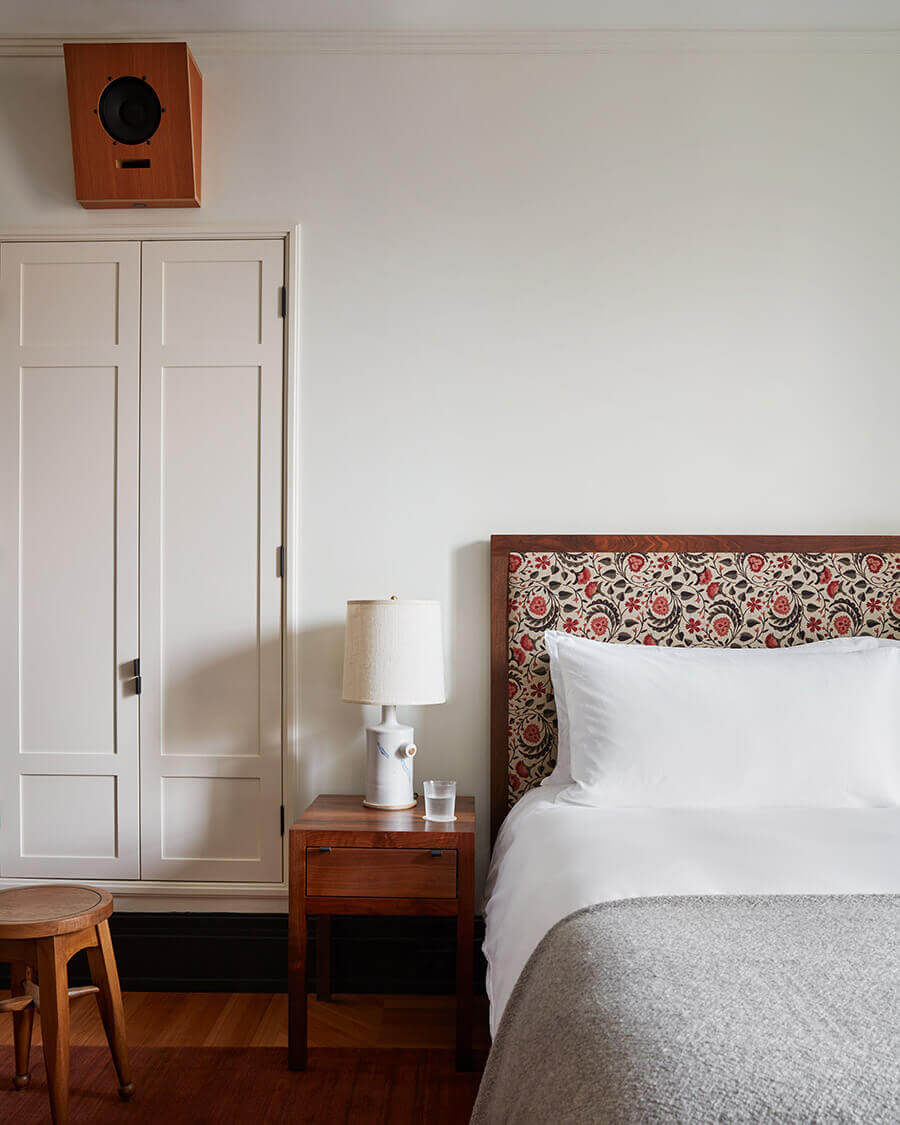 Nine Orchard, North America — Minimalist bedroom with floral patterned headboard, white linens, wooden nightstand, cream wardrobe, and geometric birdhouse detail on pale walls.