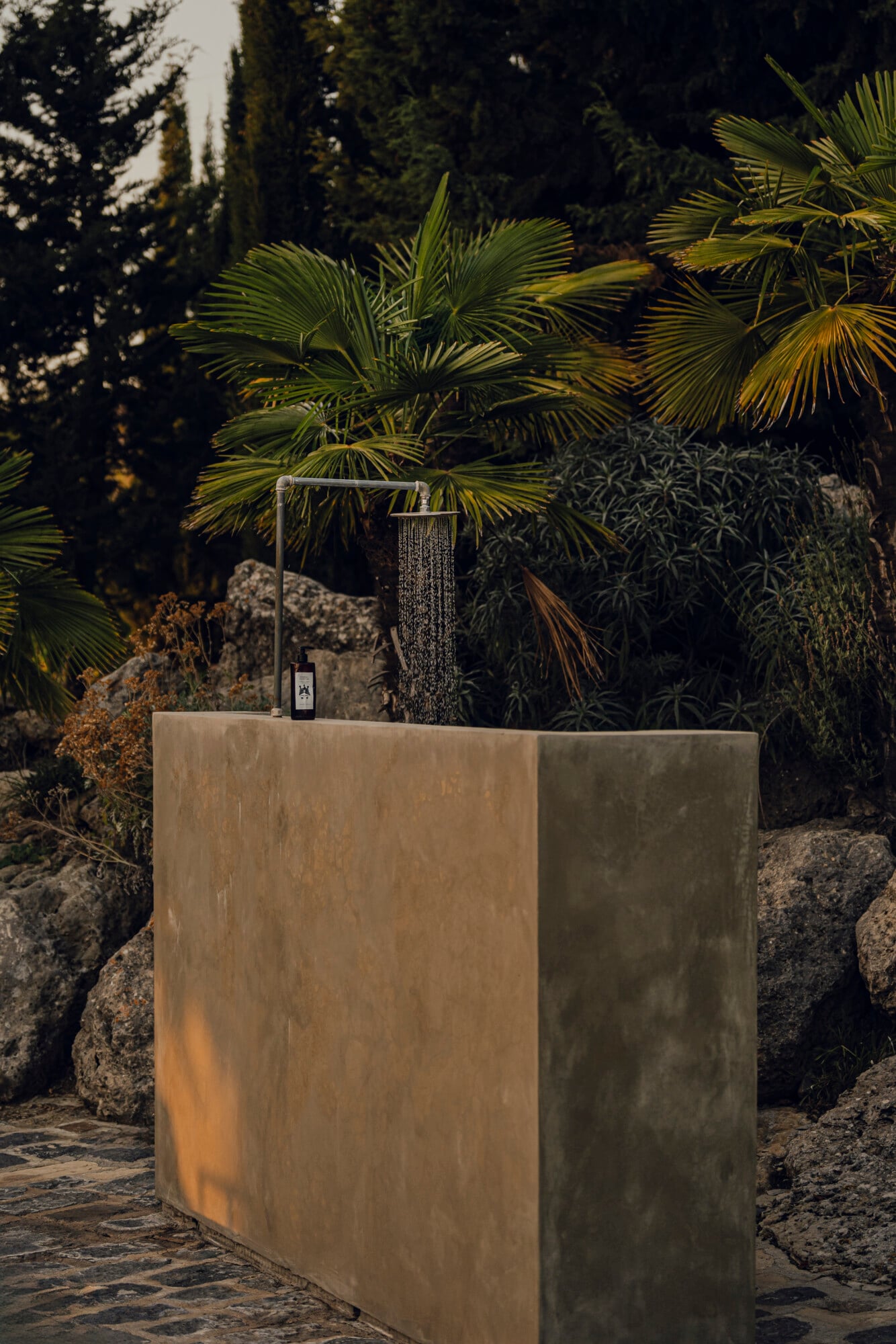 Susafa, Italy — Concrete outdoor shower with rainfall showerhead nestled among palm trees and stone walls, dappled sunlight filtering through Mediterranean foliage.