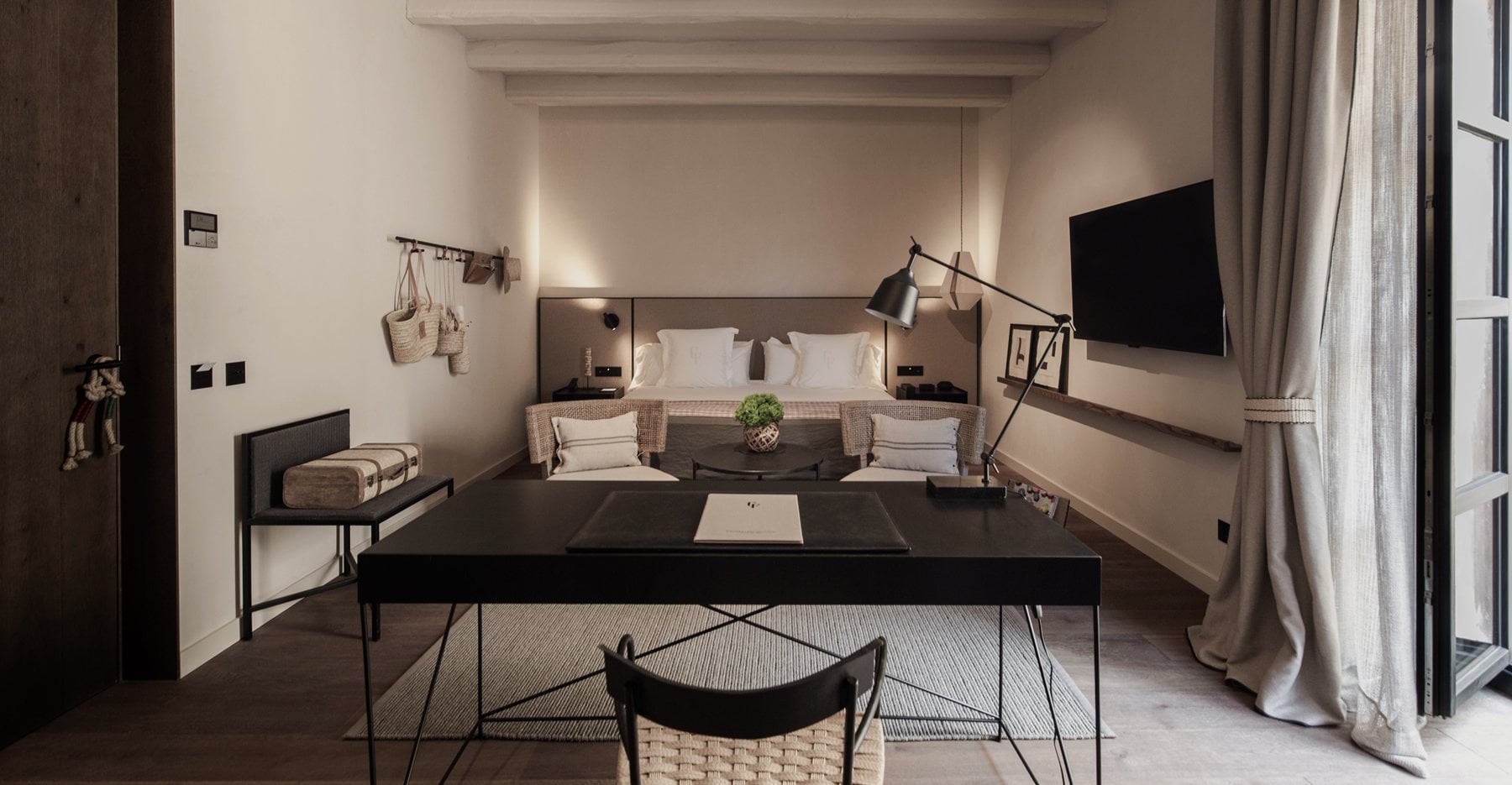 Can Ferrereta, Balearic Islands — Minimalist hotel bedroom with natural linen, black desk, woven chairs, pendant lamp, and soft north light from tall windows.