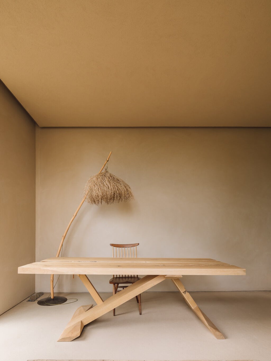 Melides Art, Portugal — Minimalist dining space with light wood table, geometric base, spindle-back chair, and sculptural dried grass floor lamp against warm neutral walls.