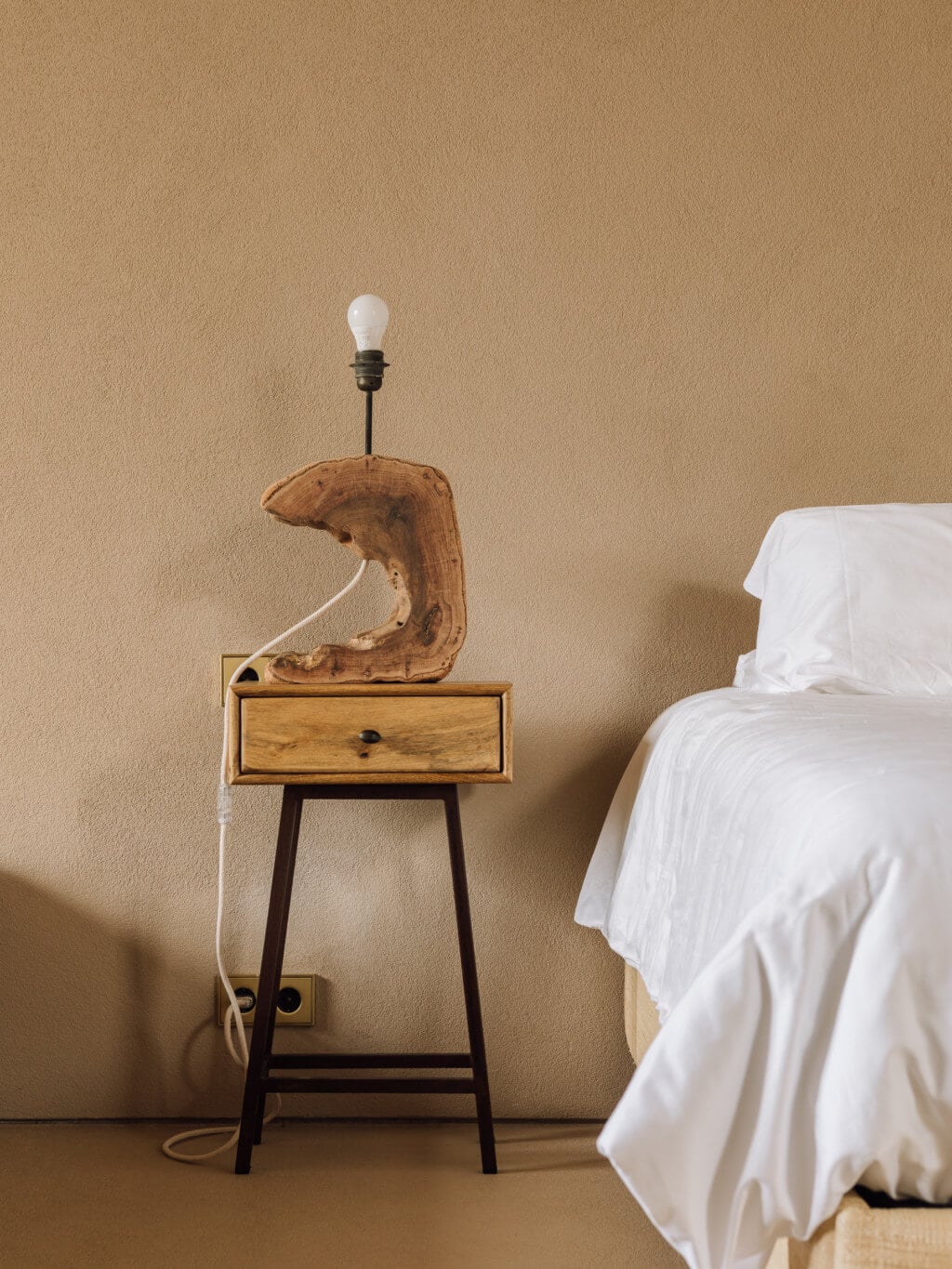 Melides Art, Portugal — Wooden sculptural lamp base with curved form on mid-century nightstand beside bed with white linens, warm neutral interior.