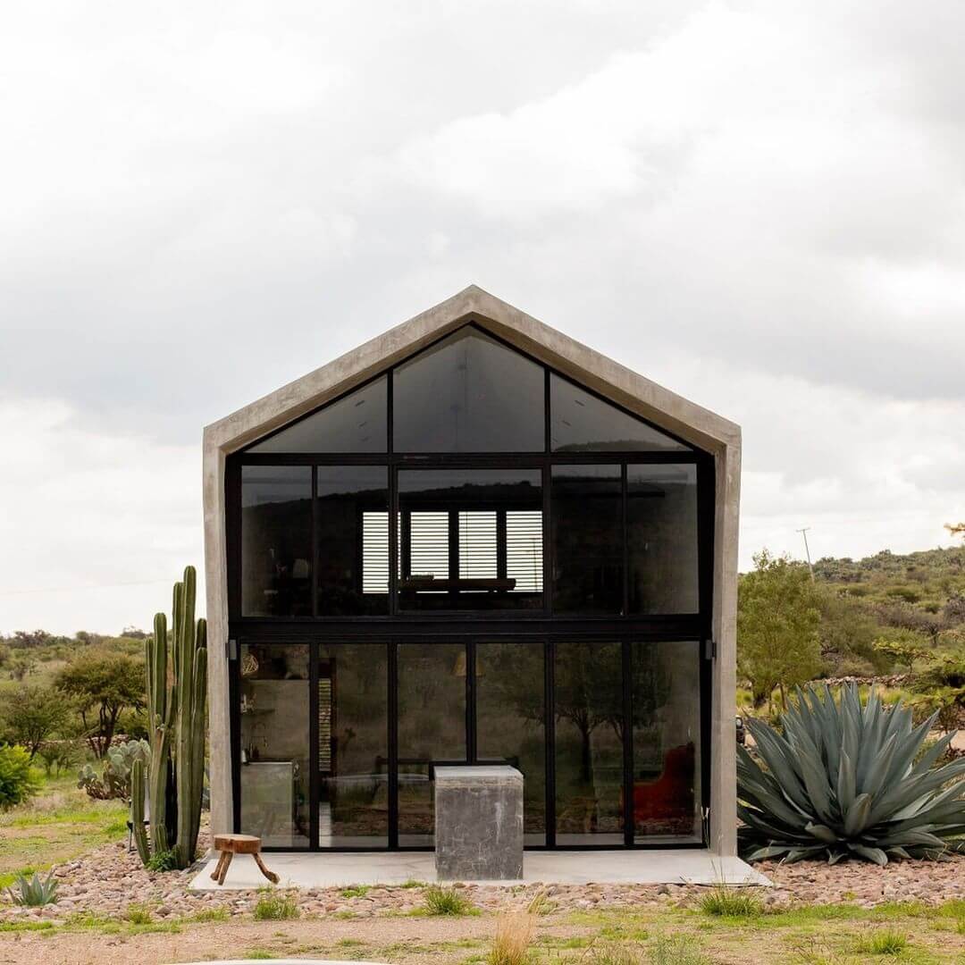 Villa ITO, Mexico — Modernist concrete gable structure with floor-to-ceiling black-framed glazing, desert landscape, agave plants, minimalist aesthetic.