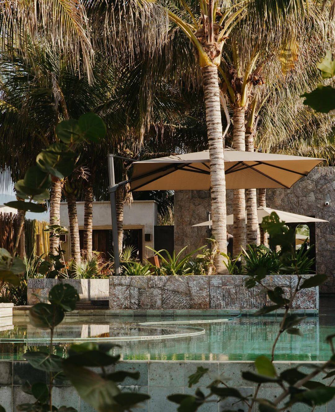Hotelito MUSA, Mexico — Minimalist poolside pavilion with tan cantilever umbrella, whitewashed walls, palm trees, and turquoise water reflecting architectural lines.
