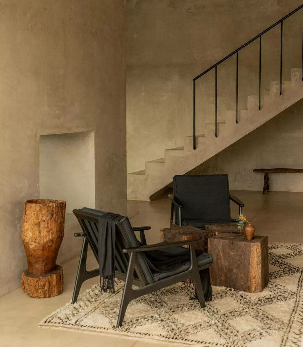 Villa ITO, Mexico — Minimalist concrete interior with black mid-century chair, wooden sculptural vessels, and floating staircase with metal railings overlooking terrazzo floor.
