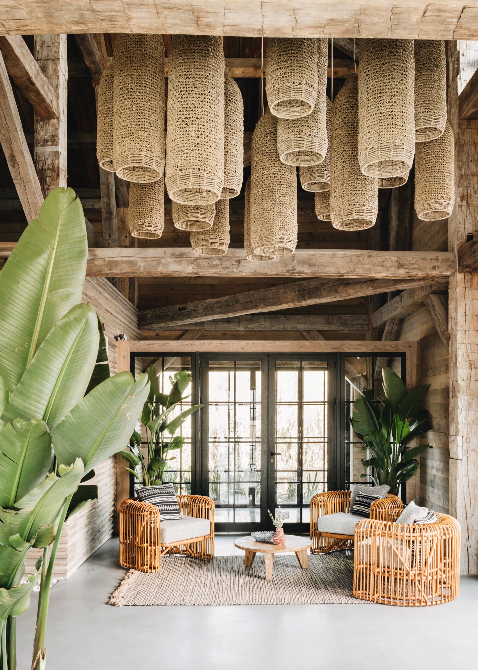 Quinta da Comporta, Europe — Woven pendant lamps hang above rattan chairs with white cushions arranged around a wooden table in a rustic-modern pavilion with polished concrete floors and black-framed glass doors.