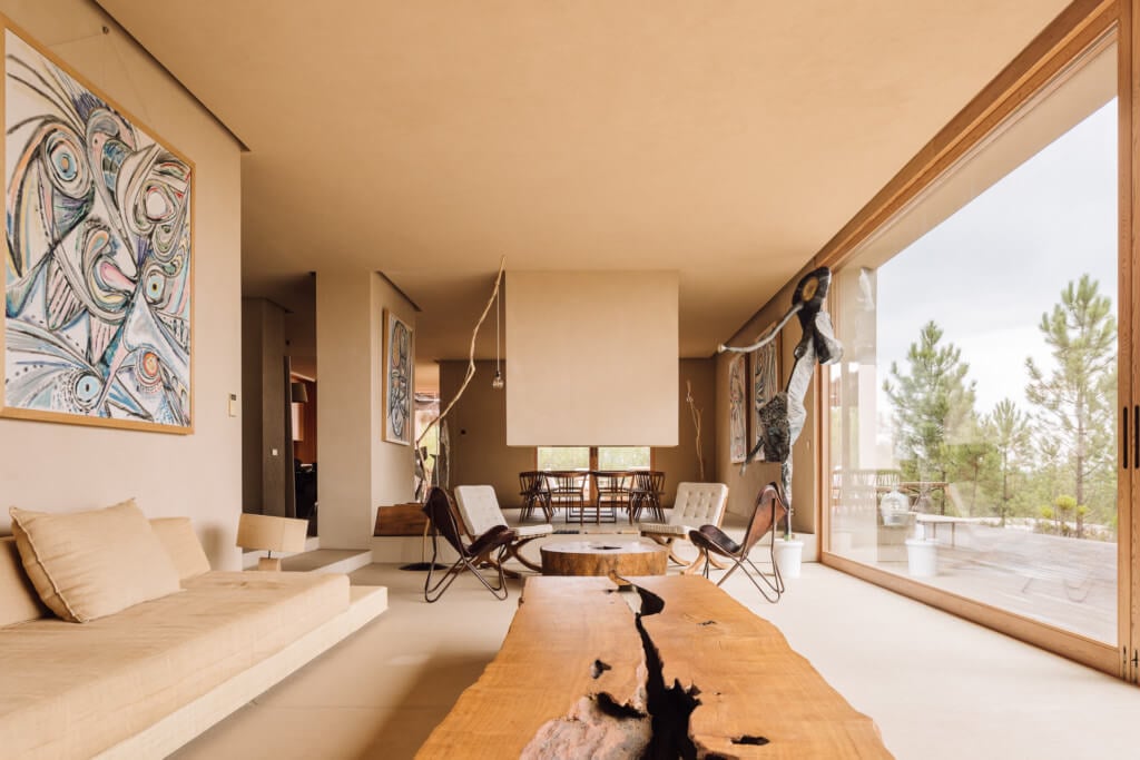 Melides Art, Portugal — Open-plan gallery living space with wooden furniture, abstract artwork, floor-to-ceiling windows overlooking pine forest, and natural light.