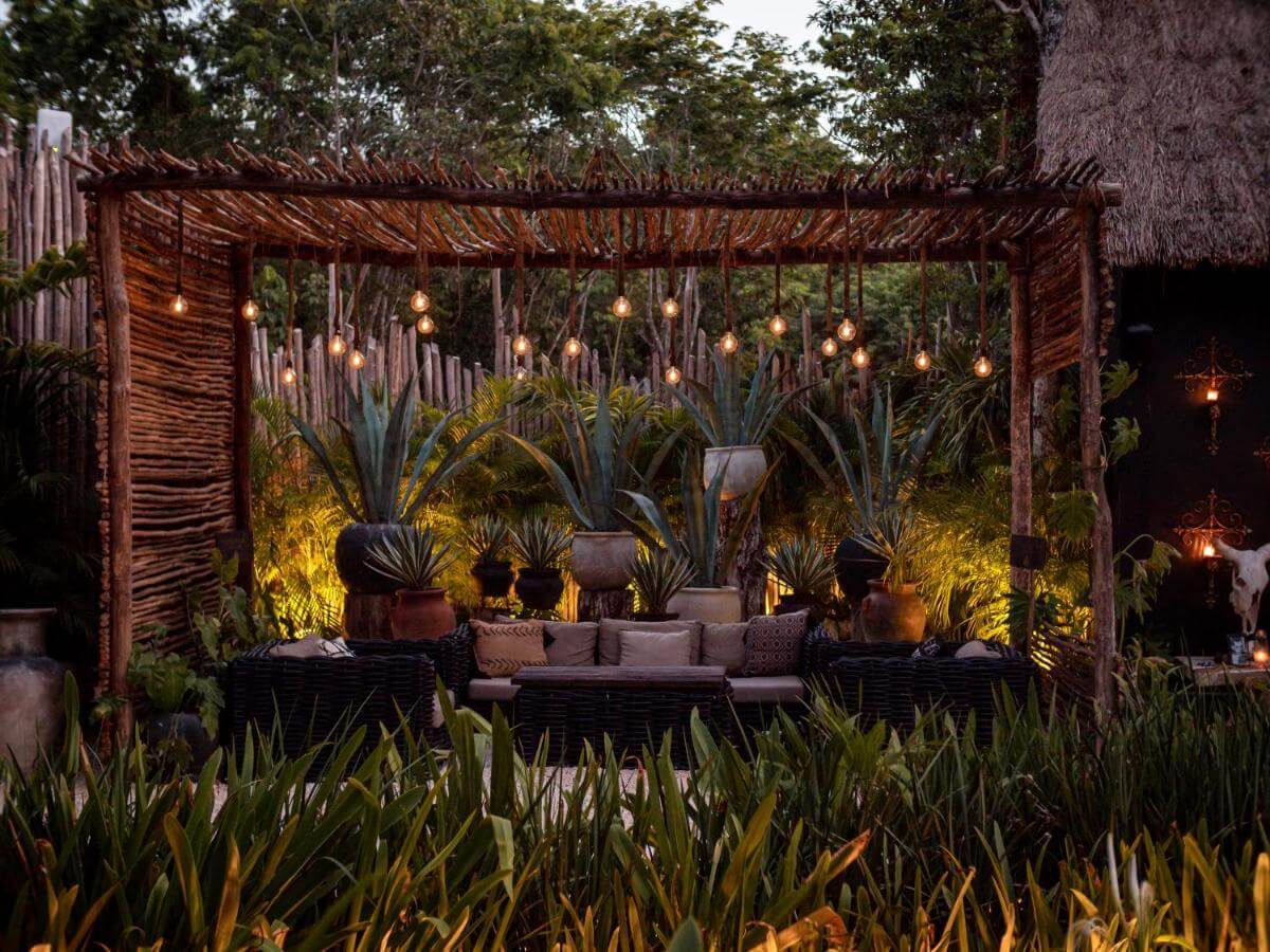 Bardo, Mexico — Wooden pergola with hanging Edison bulbs, layered uplighting on agave plants, gray cushioned seating, tropical garden at dusk.
