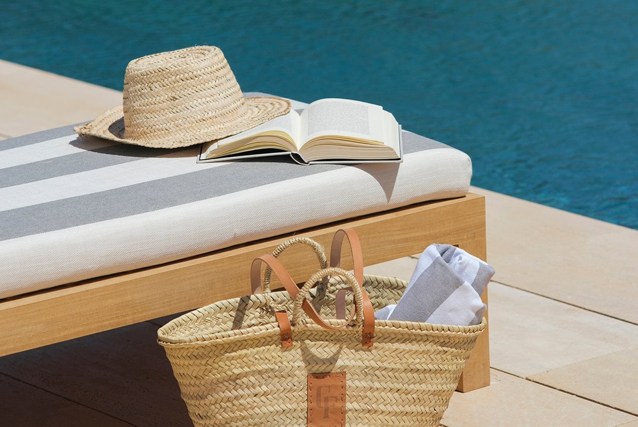 Can Ferrereta, Balearic Islands, Spain — Striped lounger with open book and straw hat beside woven basket, turquoise water beyond.