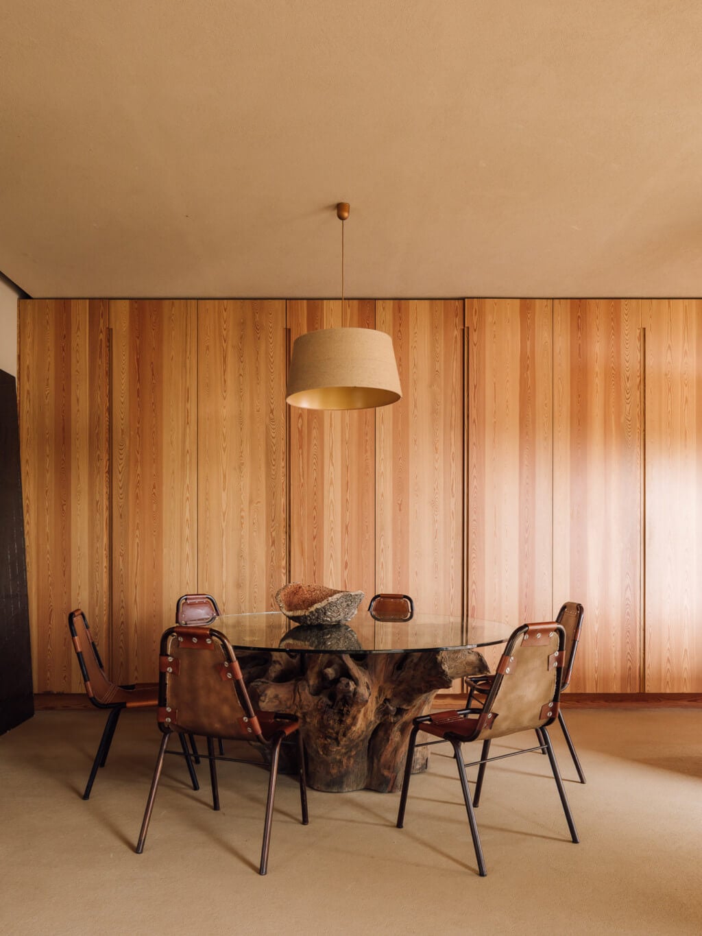 Melides Art, Portugal — Dining room with wooden wall paneling, sculptural tree-trunk base table, leather chairs, pendant lamp, and natural light.