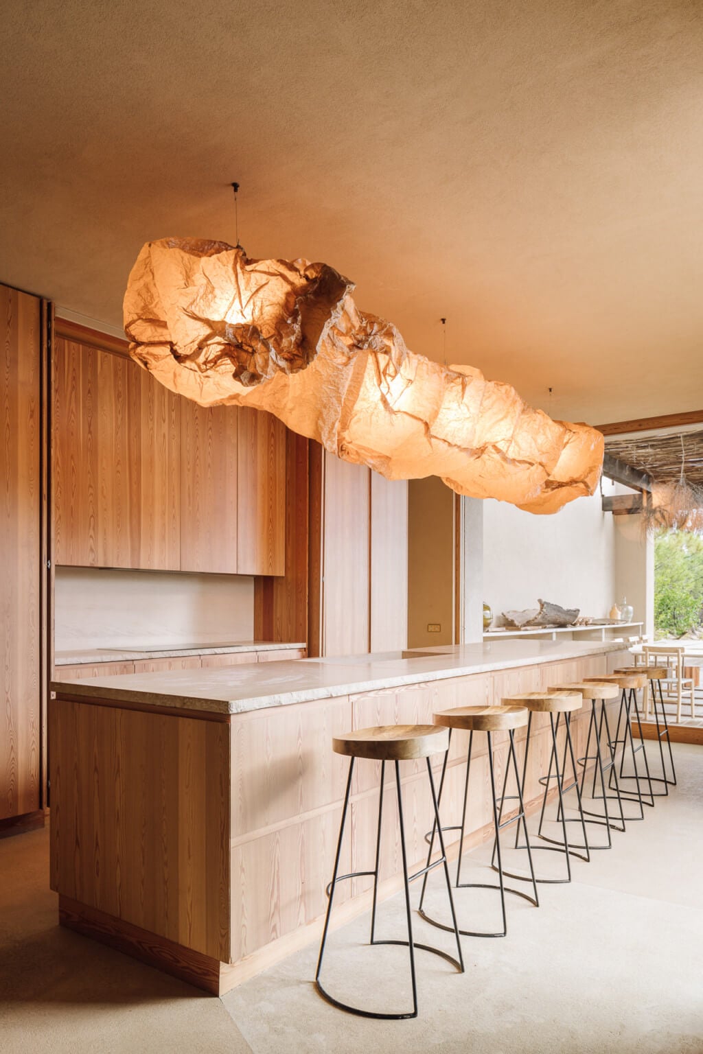 Melides Art, Portugal — Minimalist kitchen with sculptural paper pendant light, wooden cabinetry, white stone countertop, and metal bar seating in warm natural light.