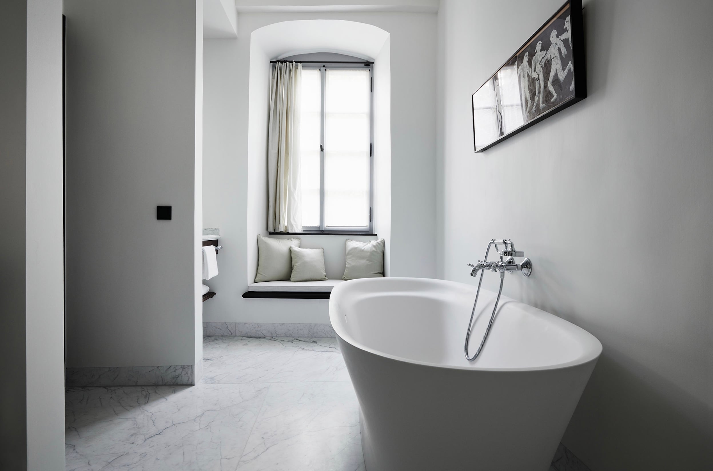 Purs, Europe — Minimalist marble bathroom with freestanding white oval bathtub, arched window with cushioned seat, and contemporary chrome fixtures.