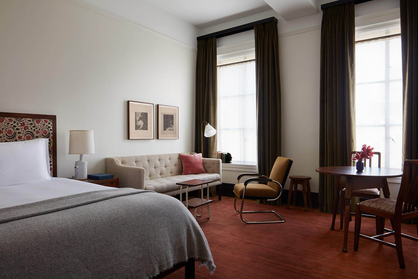 Nine Orchard, North America — Sunlit hotel suite with cream sofa, mid-century furniture, red geometric headboard, olive curtains, and warm wood accents.