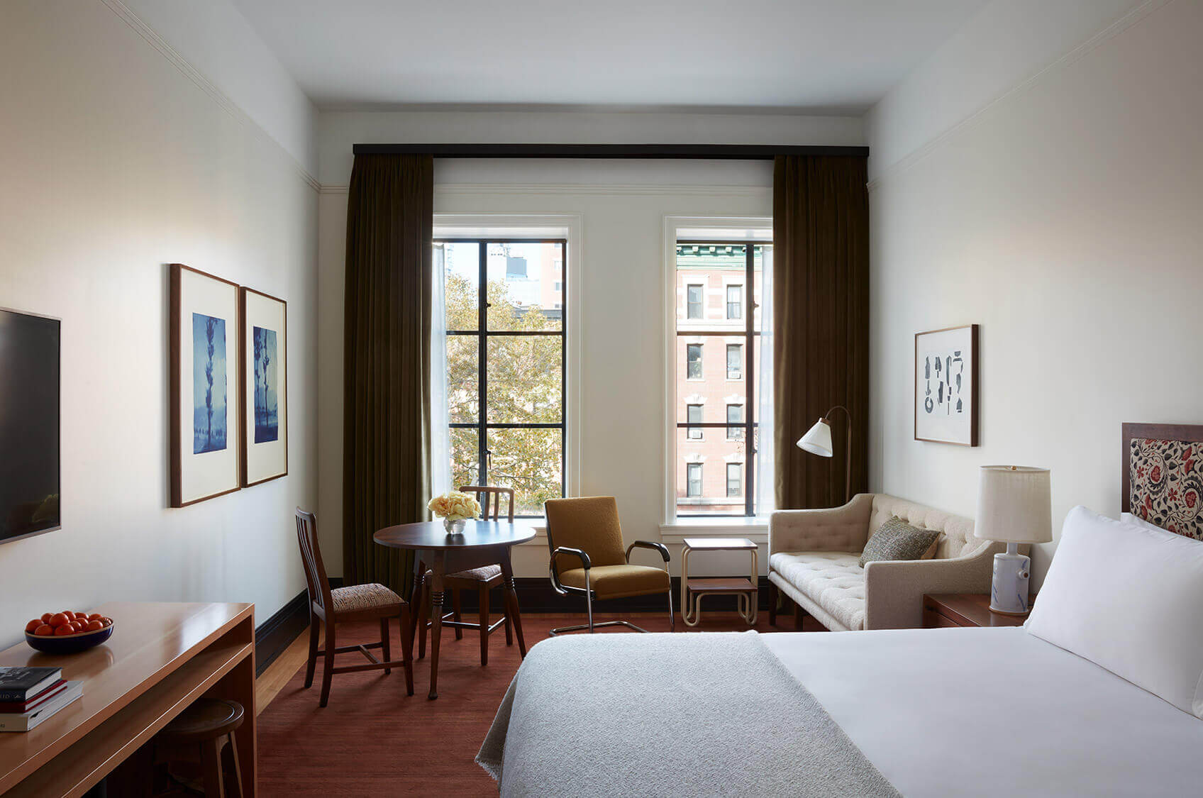Nine Orchard, North America — Modern hotel room with wood furnishings, floor-to-ceiling windows overlooking city, brown curtains, cream bedding, mid-century seating area, warm minimalist aesthetic.