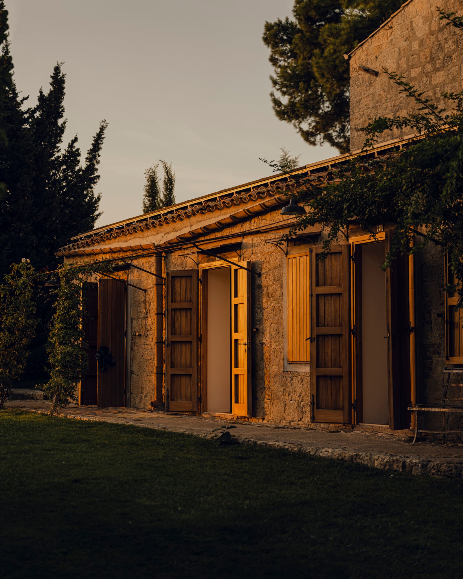 Susafa, Italy — Stone cottage with golden-hour light, wooden doors and pergola, cypress trees, rustic Mediterranean architecture and manicured grounds.