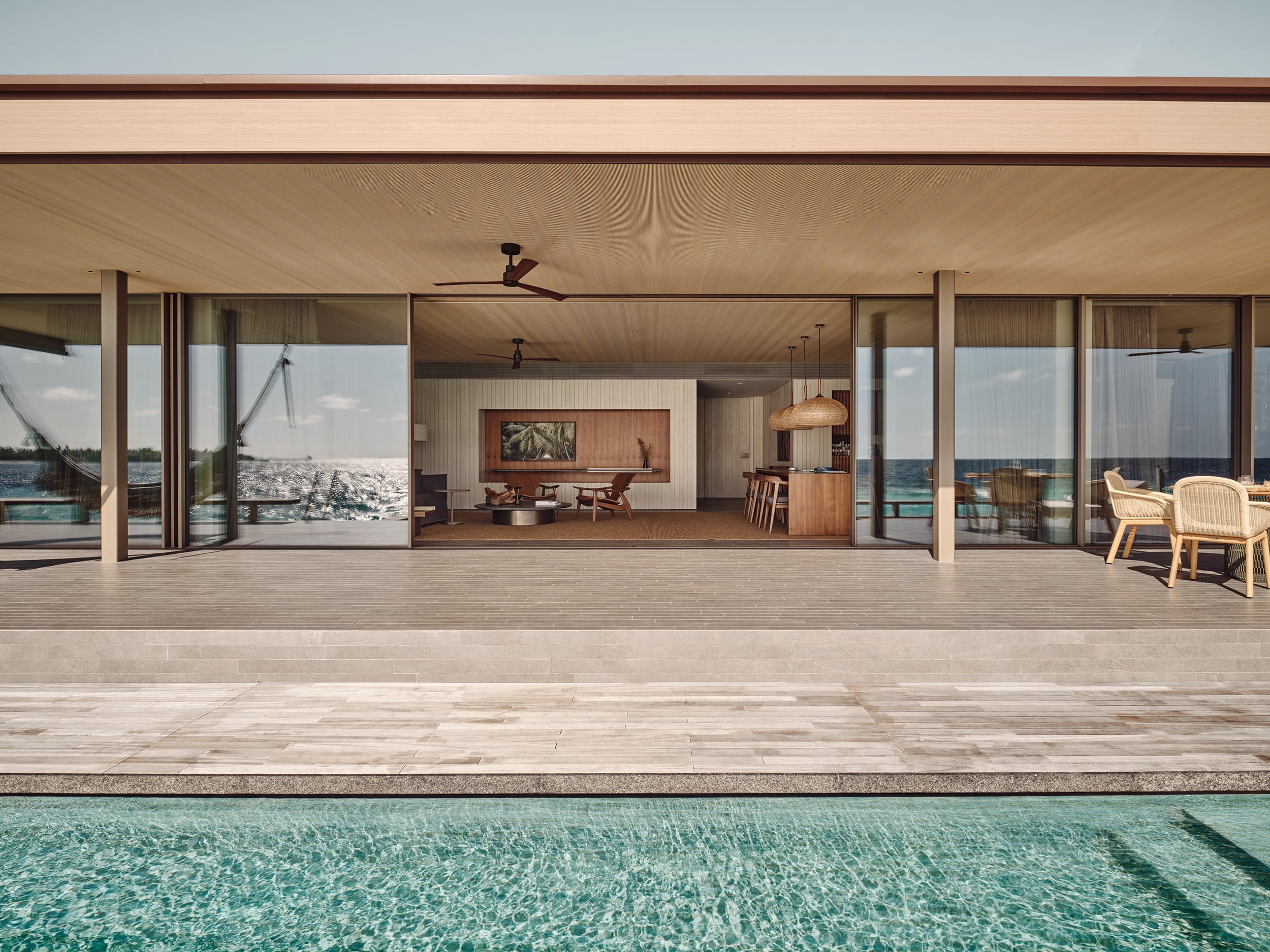Patina Maldives, Asia — Modern waterfront villa with floor-to-ceiling glass, wooden deck, infinity pool, and minimalist interior with ocean views.