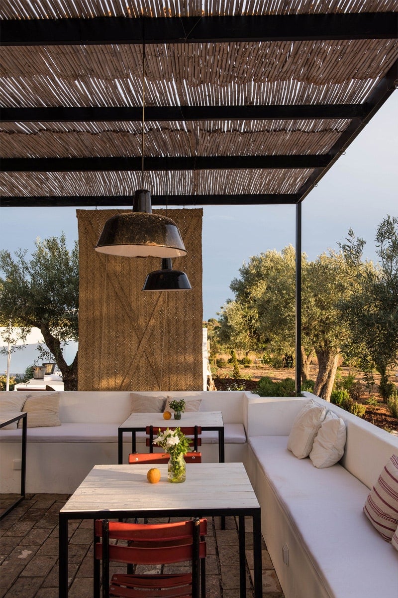 Pensão Agrícola, Europe — Shaded terrace with woven timber pergola, white built-in seating, industrial pendant lights, olive grove views, Mediterranean hospitality design.