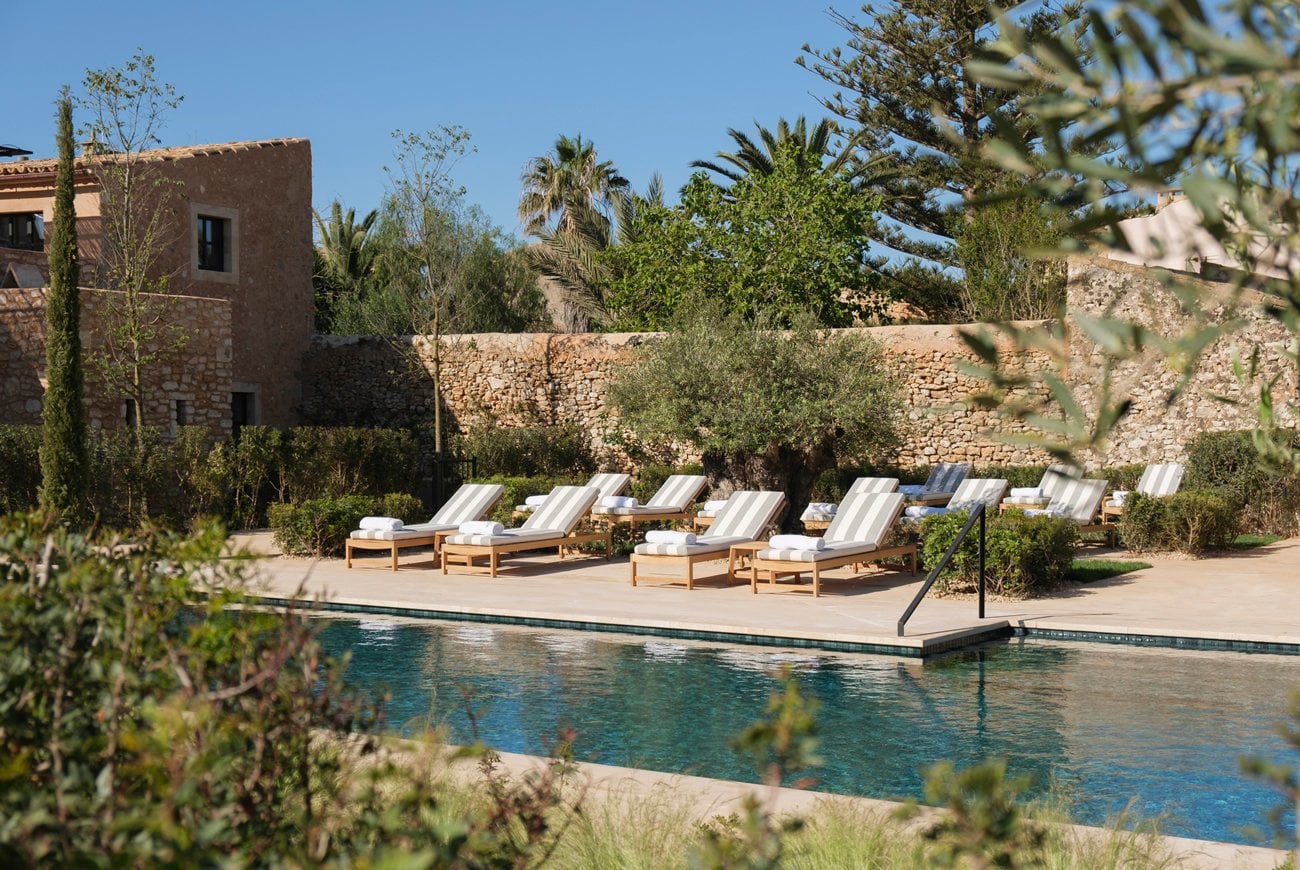 Can Ferrereta, Balearic Islands, Spain — Poolside lounge chairs with striped cushions, terracotta architecture, olive trees, and stone walls under clear sky.