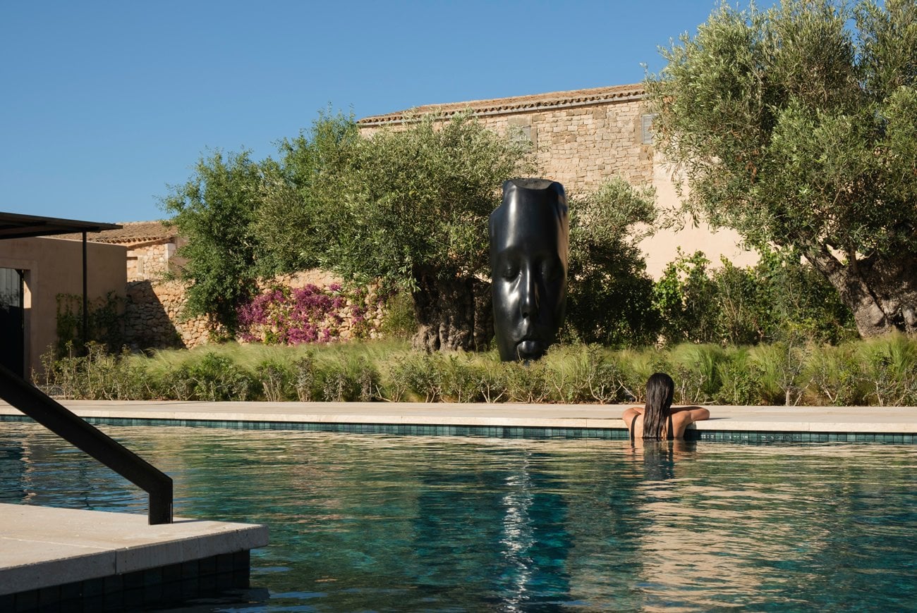 Can Ferrereta, Balearic Islands — Woman swimming in turquoise pool with black sculptural art, stone architecture, olive trees and magenta bougainvillea under clear sky.