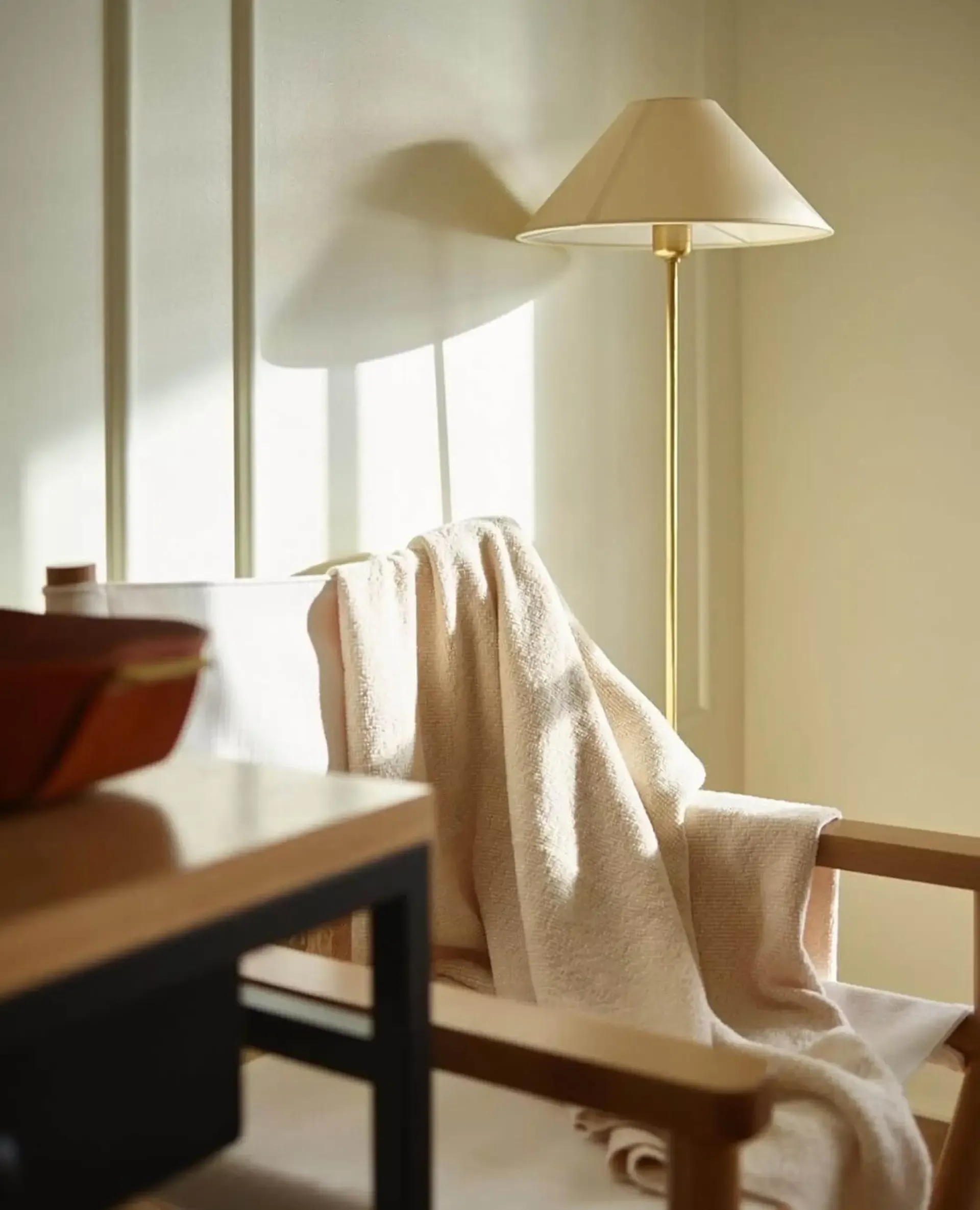 The Lake House, North America — Cream linen throw draped over wooden chair beside brass floor lamp with cone shade, bright windows casting soft shadows on warm walls.