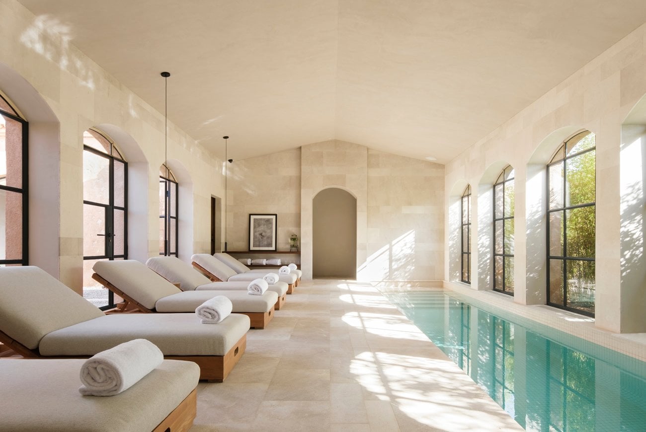 Sa Calma Spa, Balearic Islands, Spain — Indoor lap pool with cream stone walls, wooden loungers with rolled towels, arched doorway, and natural light through tall windows.
