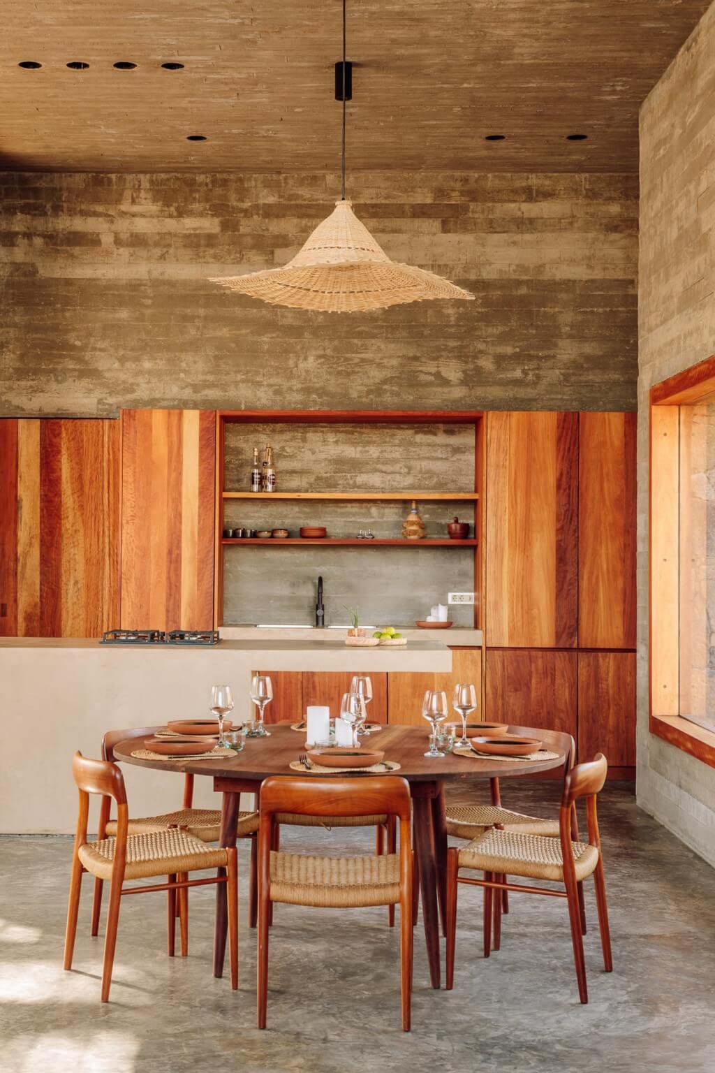 Barefoot Luxury, Africa — Dining room with round wood table, woven chairs, timber cabinetry, concrete walls, and pendant light fixture.