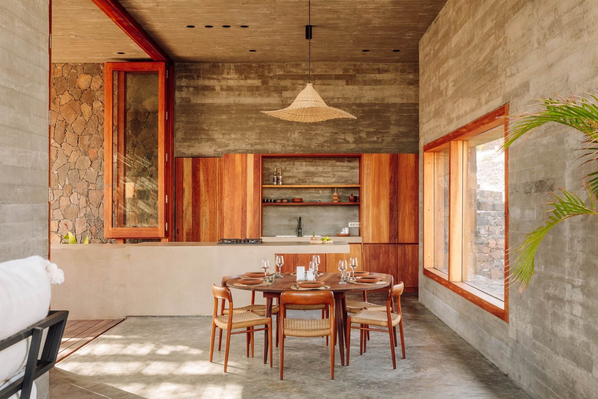 Barefoot Luxury, Africa — Open-plan dining pavilion with wooden round table, rattan chairs, concrete walls, timber-framed windows, and woven pendant light.