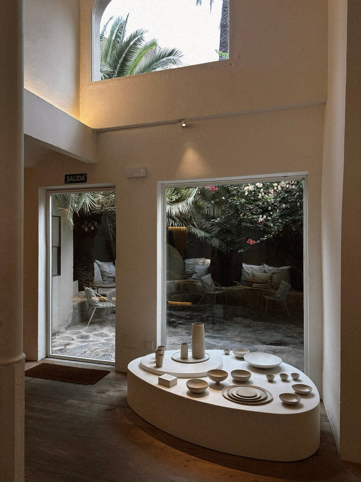 Cortana, Balearic Islands — Minimalist ceramic display in bright gallery space with high clerestory window, palm trees visible, opening to courtyard seating area.