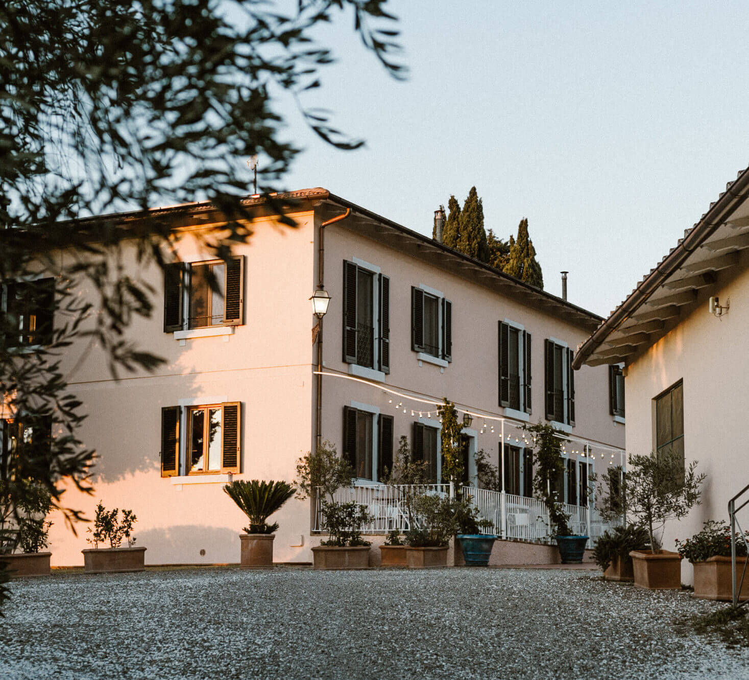 Villa Lena, Europe — Cream-colored Italian villa with black shutters, cypress trees, terracotta planters, golden hour light, gravel courtyard.