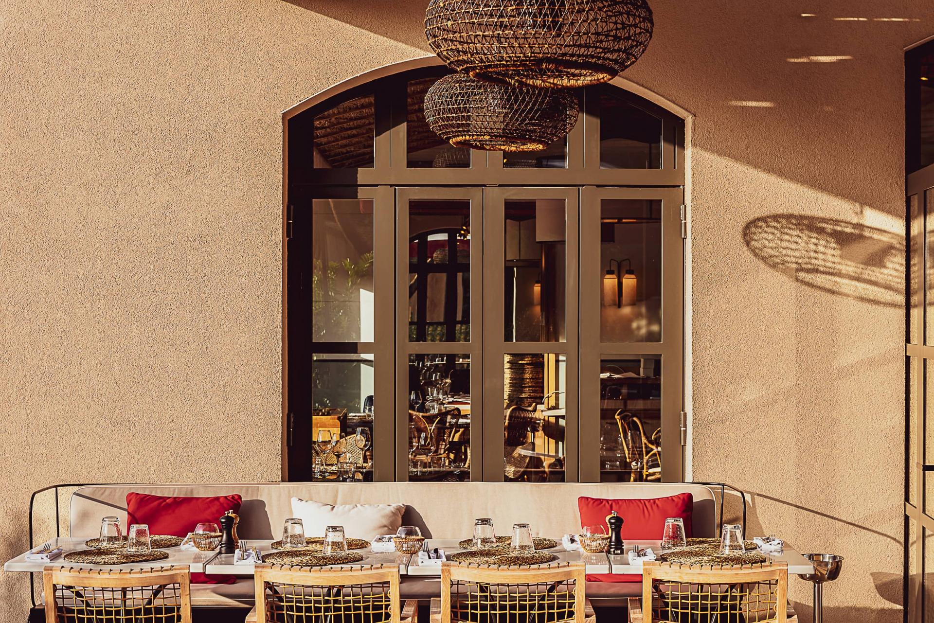 Hôtel Lou Pinet, Europe — Dining terrace with woven pendant lights, cream banquette, rattan chairs, and arched glass doors casting geometric shadows on textured walls.