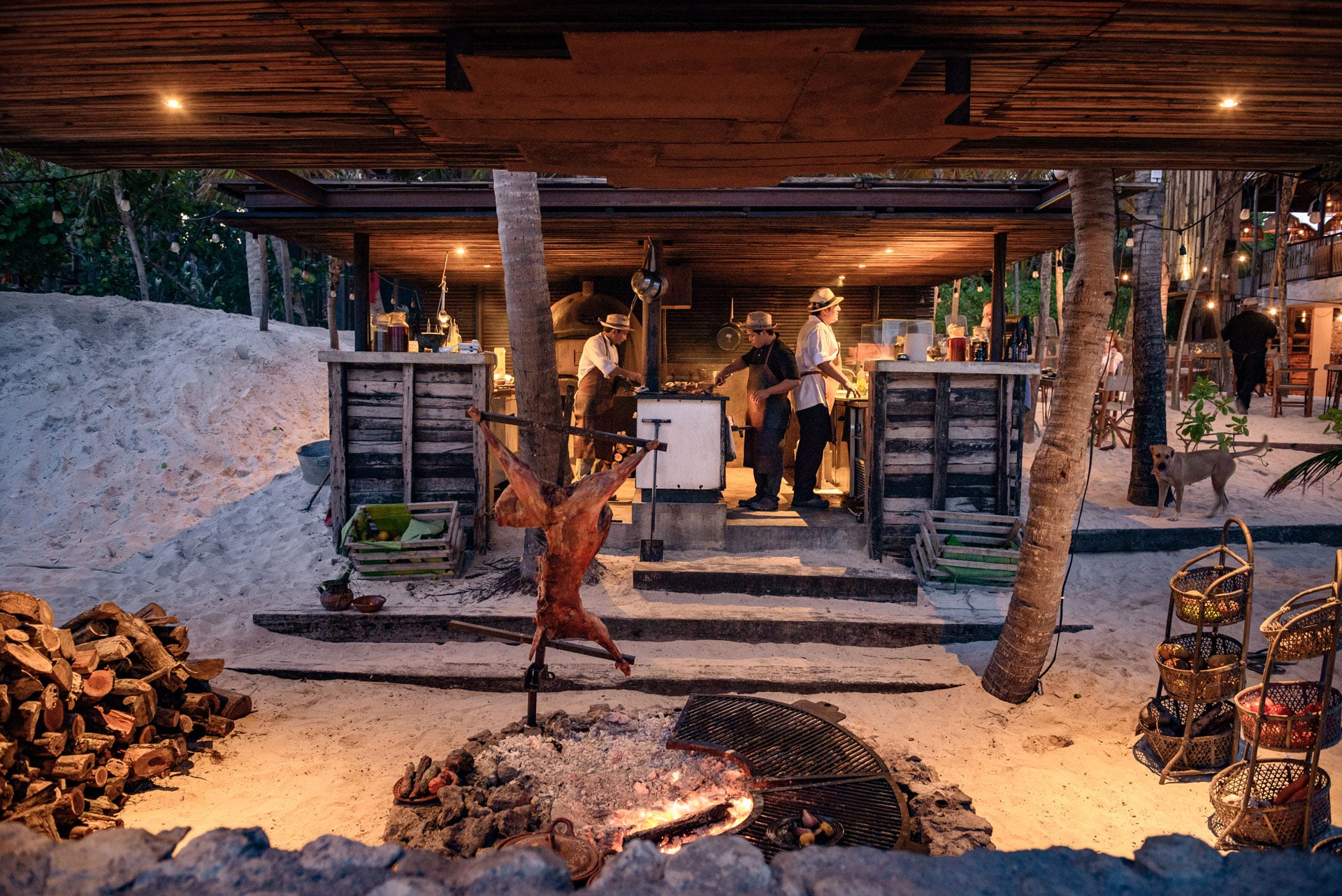 Be Tulum, Mexico — Beach bonfire gathering under wooden pergola at dusk, rustic grill and woven baskets, warm ambient lighting.
