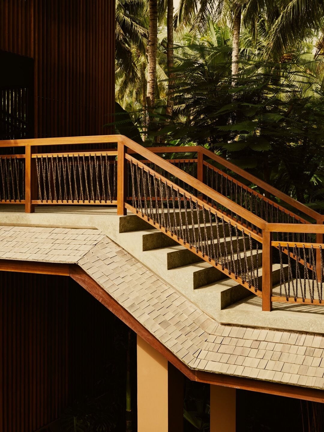 Lost Lindenberg, Asia — Wooden deck with cable railing overlooking tropical gardens, afternoon light casting shadows across composite steps and pale stone terrace.