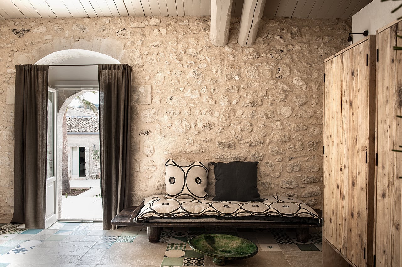 Dimora delle Balze, Europe — Stone bedroom with arched window, dark daybed, geometric pillows, wooden doors, and traditional tile flooring.