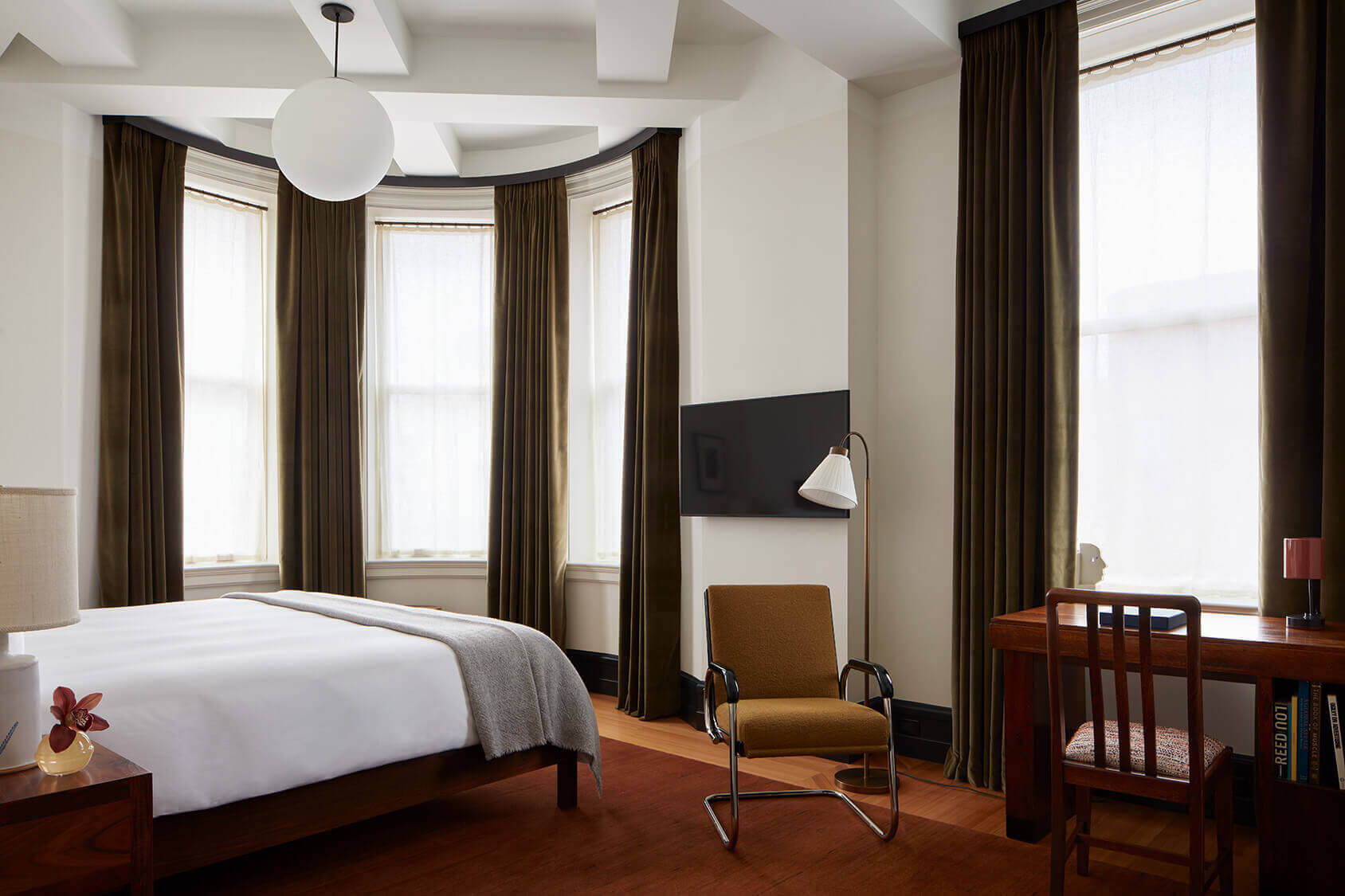 Nine Orchard, New York, USA — Luxury hotel bedroom with bay windows, olive curtains, mid-century modern furnishings, hardwood floors, and minimalist pendant lighting.