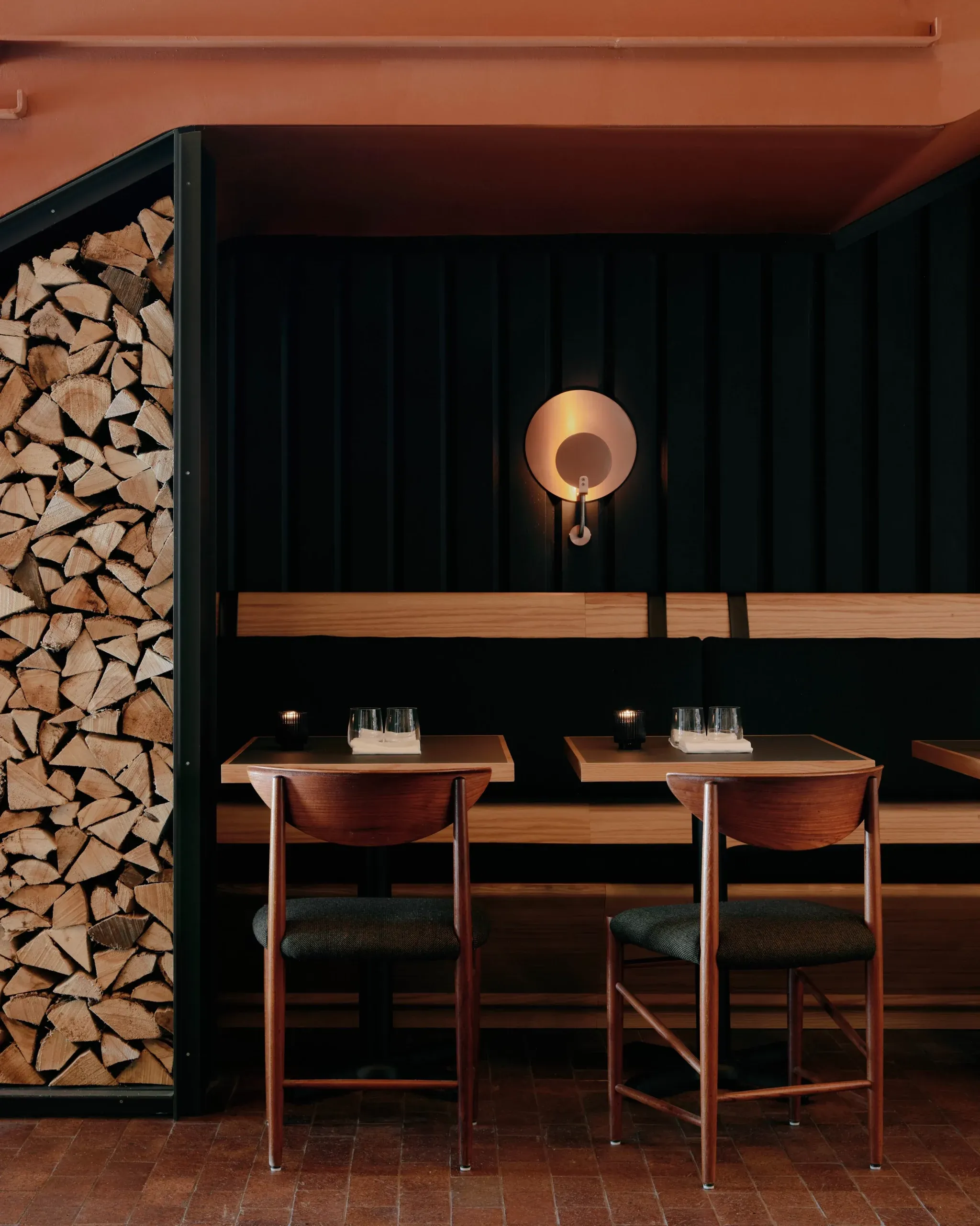 Ace Hotel Toronto, Canada — Mid-century wooden chairs at minimalist bar counter with copper pendant light, dark corrugated wall, and stacked firewood detail.