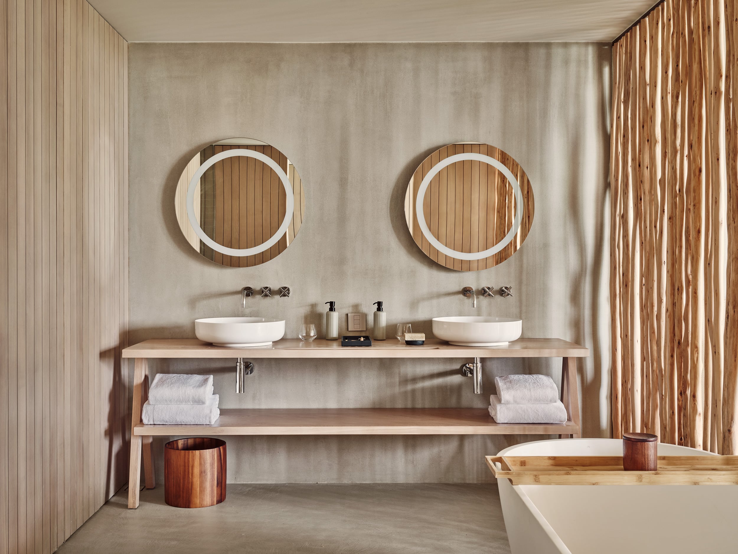 Patina Maldives, Asia — Spa bathroom with dual vessel sinks, circular mirrors with wooden frames, natural light, and minimalist wooden shelving in neutral tones.
