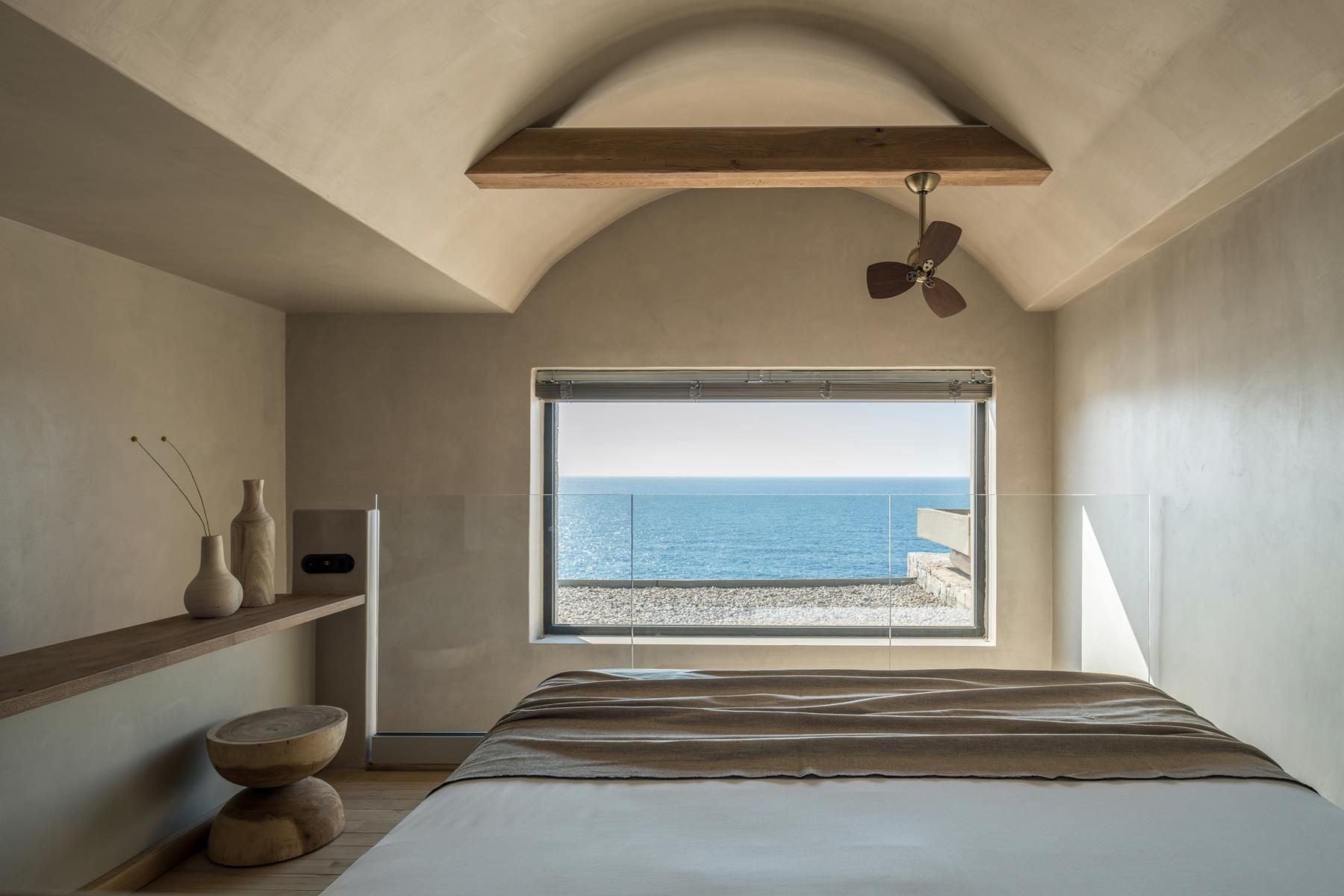 ACRO Suites, Europe — Minimalist beachfront bedroom with curved ceiling, wooden beam, sea-view window, natural materials, and small fan.