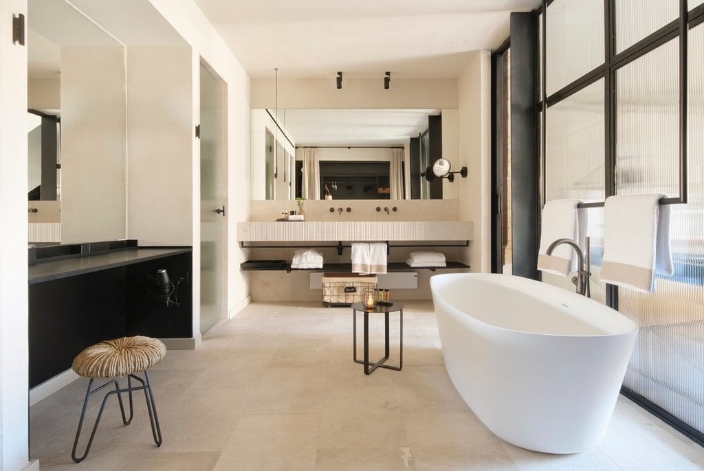 Can Ferrereta, Balearic Islands — Minimalist bathroom with white freestanding tub, dual vanity, black accents, natural stone flooring, and abundant natural light.