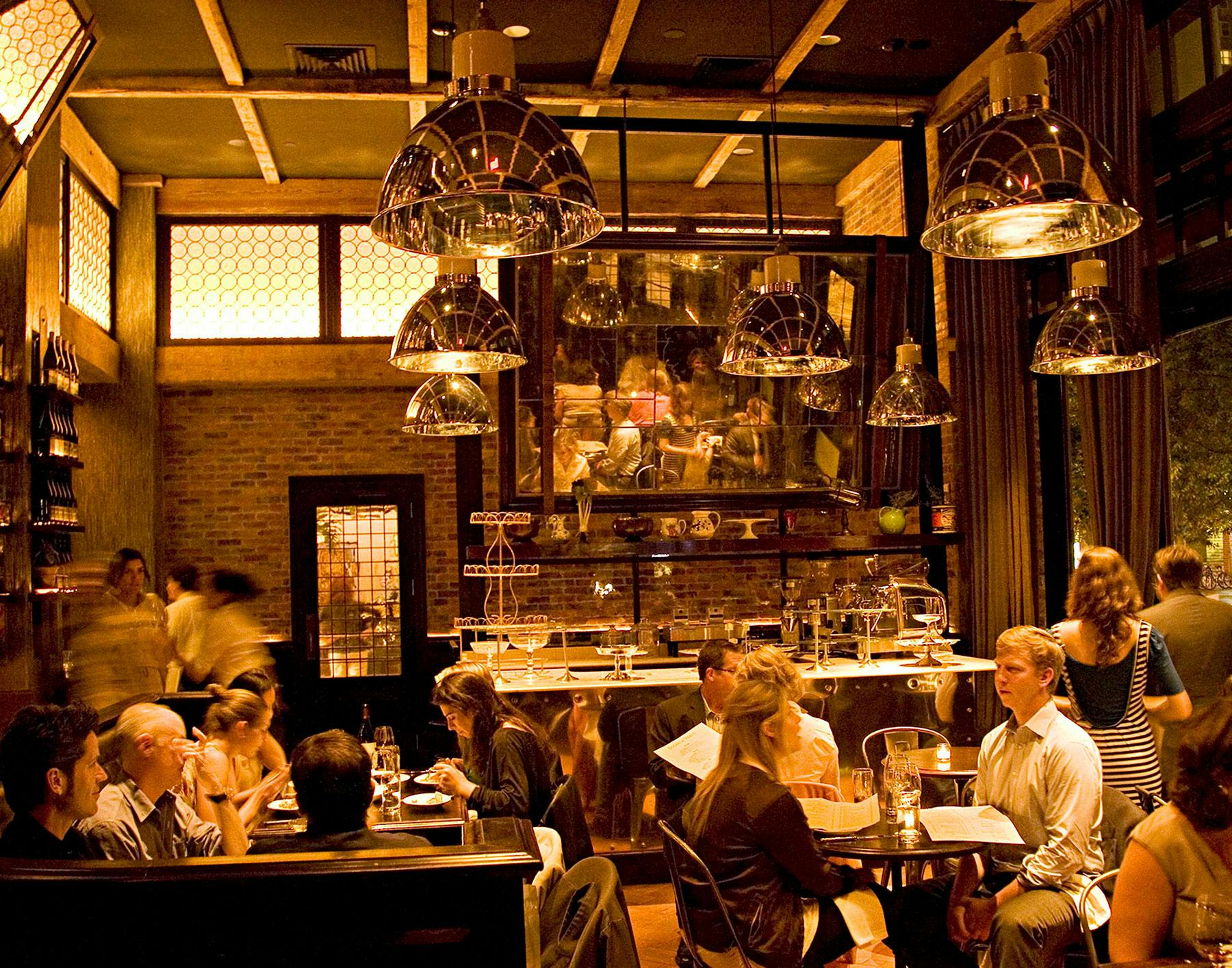 Locanda Verde, North America — Warm-lit open kitchen with brass dome pendants, brick walls, and communal dining atmosphere.