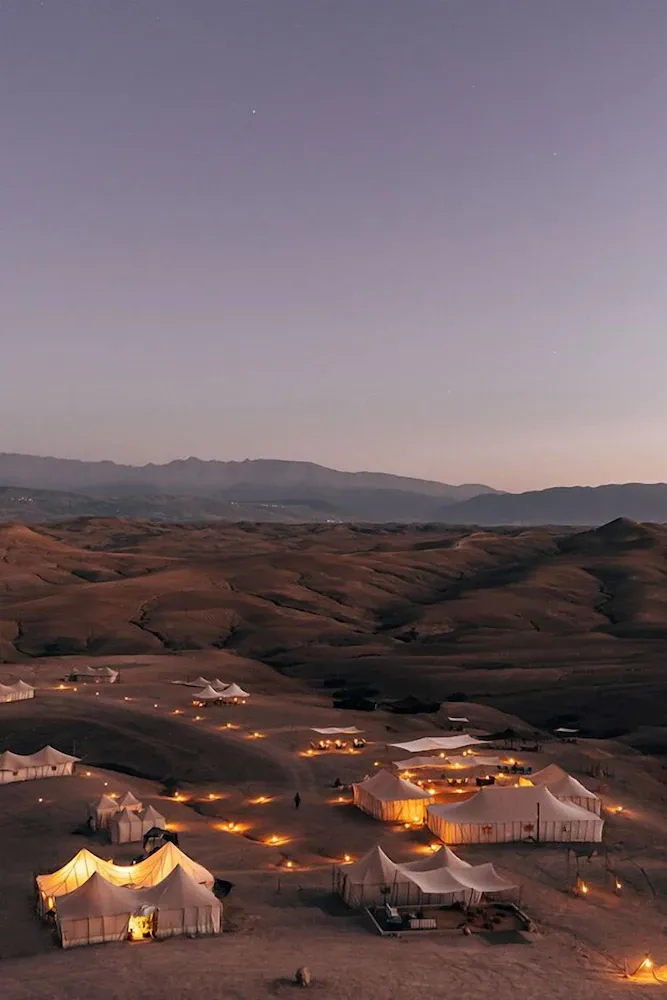 Scarabeo Camp, Africa — Illuminated Bedouin tents nestled in desert dunes at dusk, warm golden lighting casting long shadows across sand, mountains silhouetted against twilight sky.
