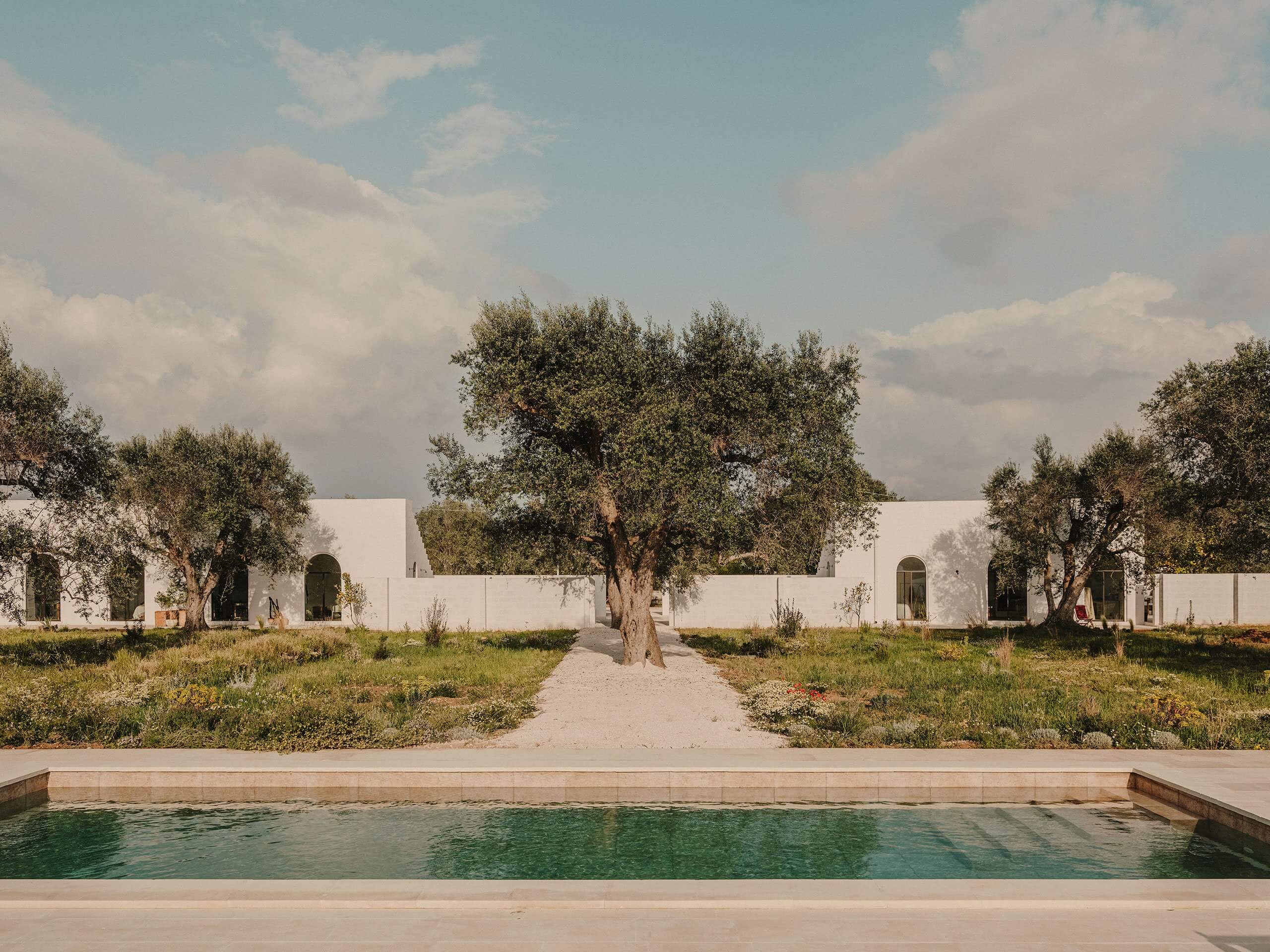 Borgo Gallana, Europe — Minimalist white courtyard pavilions with olive trees, pool edge, and gravel pathways under Mediterranean sky.