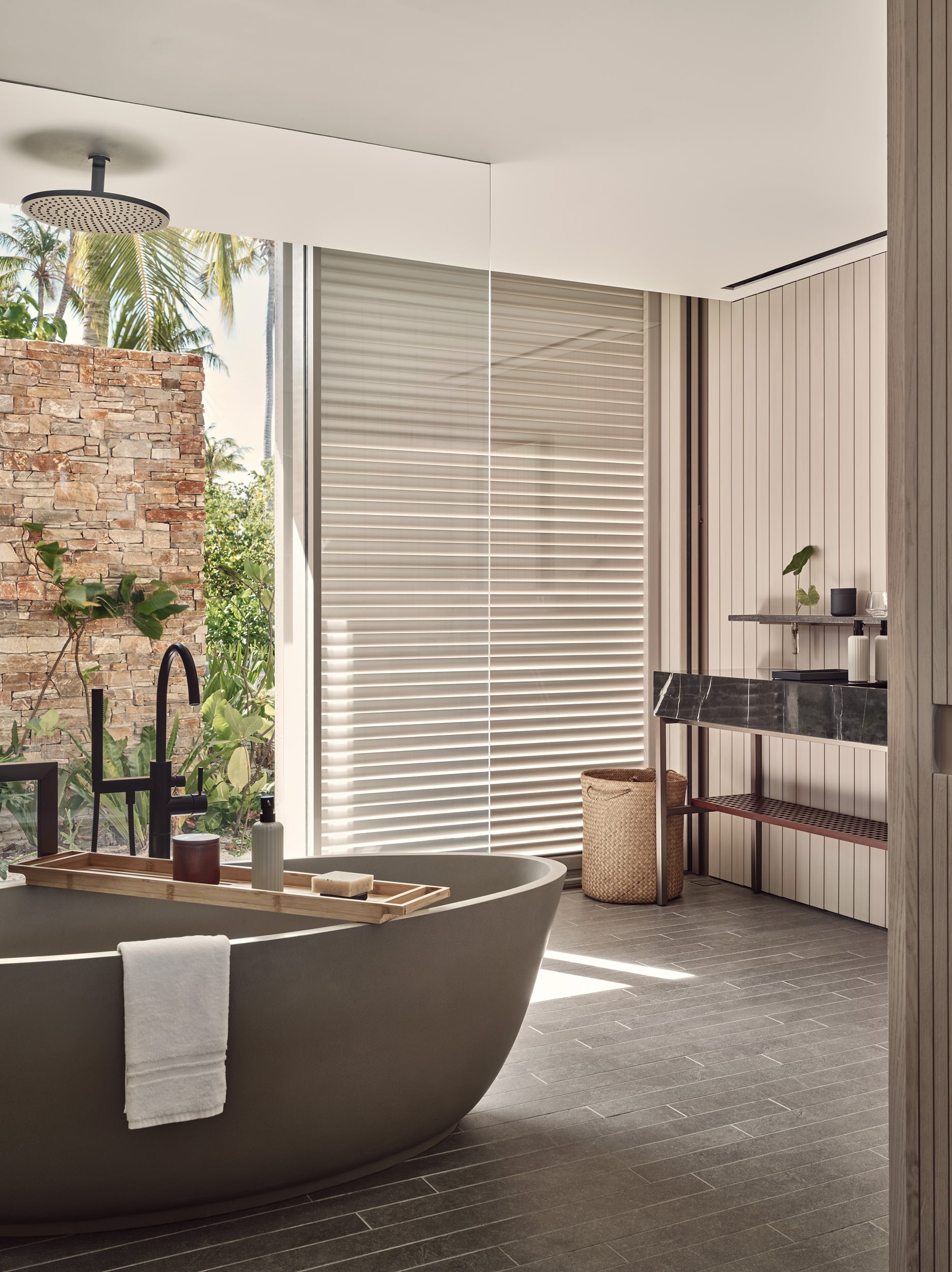 Patina Maldives, Asia — Modern spa bathroom with charcoal soaking tub, rainfall showerhead, louvered windows, stone flooring, and ocean-facing garden view.