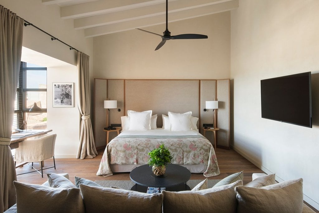 Can Ferrereta, Balearic Islands — Spacious bedroom with upholstered headboard, linen seating area, black ceiling fan, and flat-screen TV overlooking Mediterranean terrace.