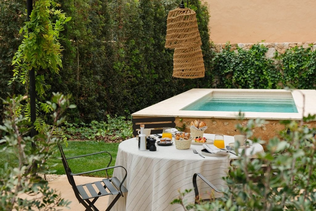 Can Ferrereta, Balearic Islands — Poolside breakfast table with woven pendant light, stone terrace, lush hedges, turquoise water.